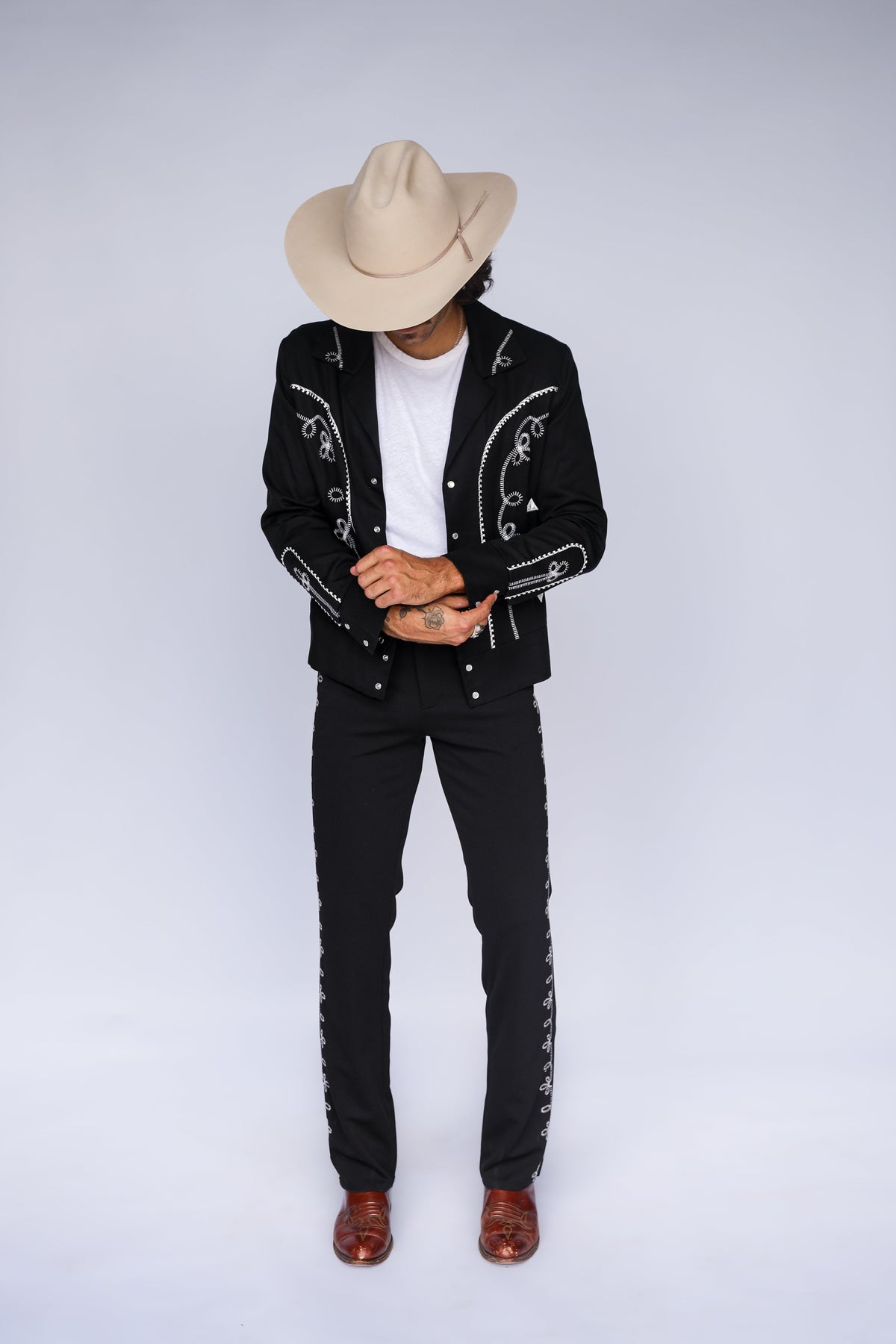 A man wearing a black western-style embroidered jacket and pants with silver trim, a beige cowboy hat, a white t-shirt, and brown boots, standing against a light gray studio background.