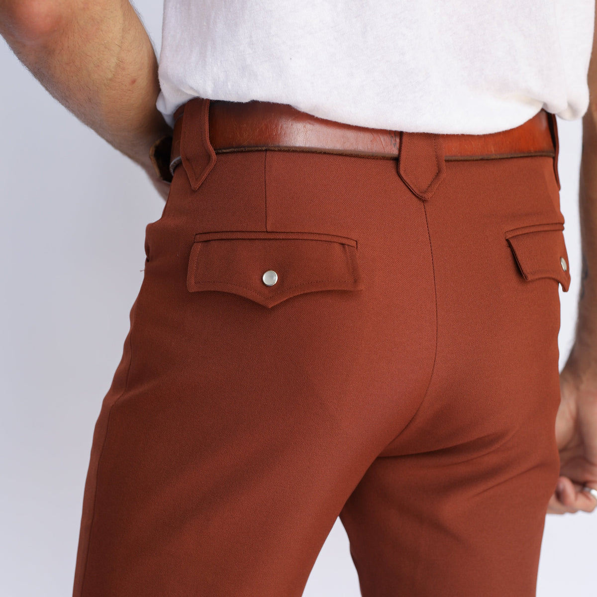 Rust-colored chinos shown from the back on a model, featuring a white t-shirt, brown leather belt, and back pocket details against a light gray background.