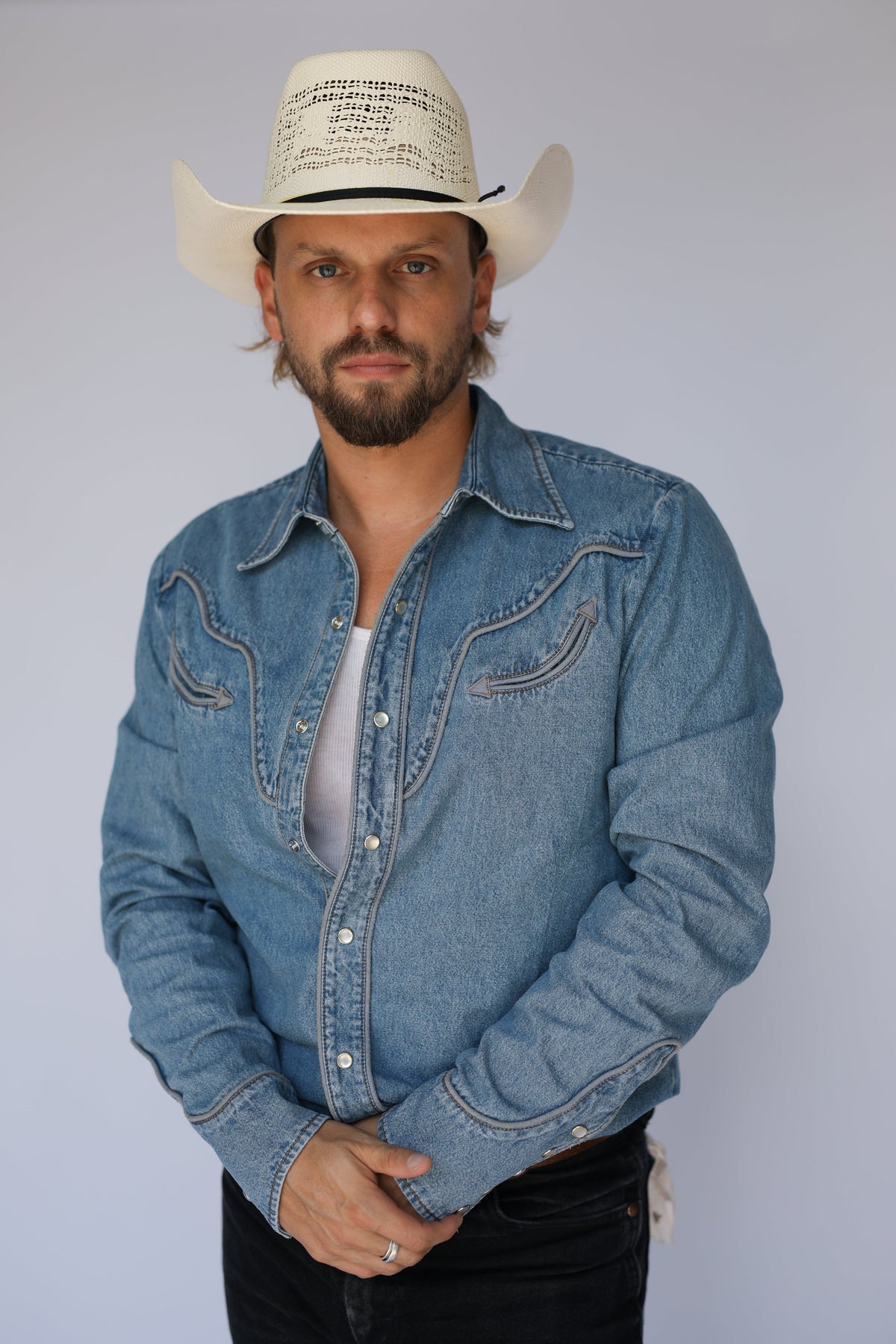 A man wearing a light blue denim western-style button-up shirt, a white cowboy hat, and black pants, standing against a plain light gray studio background.