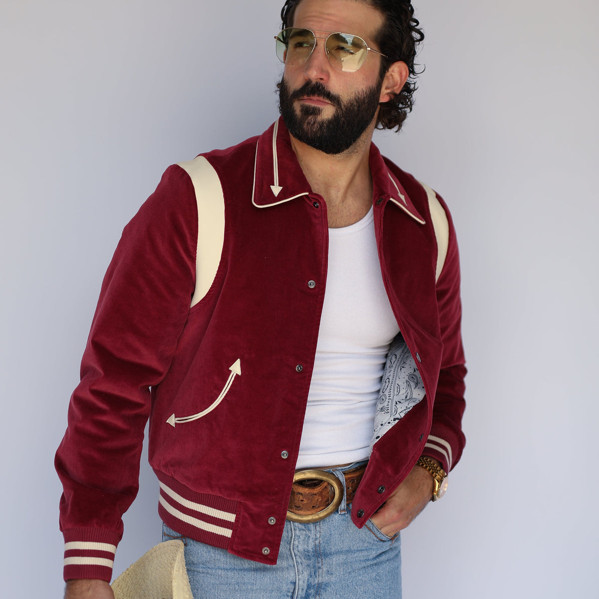 A man wearing a maroon velvet varsity-style jacket with cream ribbed trim, a white tank top, light blue jeans, and holding a cream cowboy hat, posed against a light gray studio background.