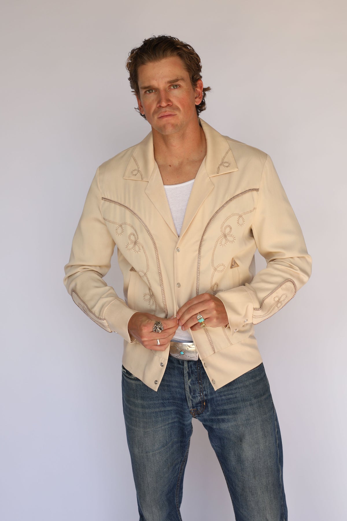A man wearing a cream H Bar C vintage inspired western-style jacket with embroidered details, a white undershirt, and blue jeans, standing against a plain light gray background.