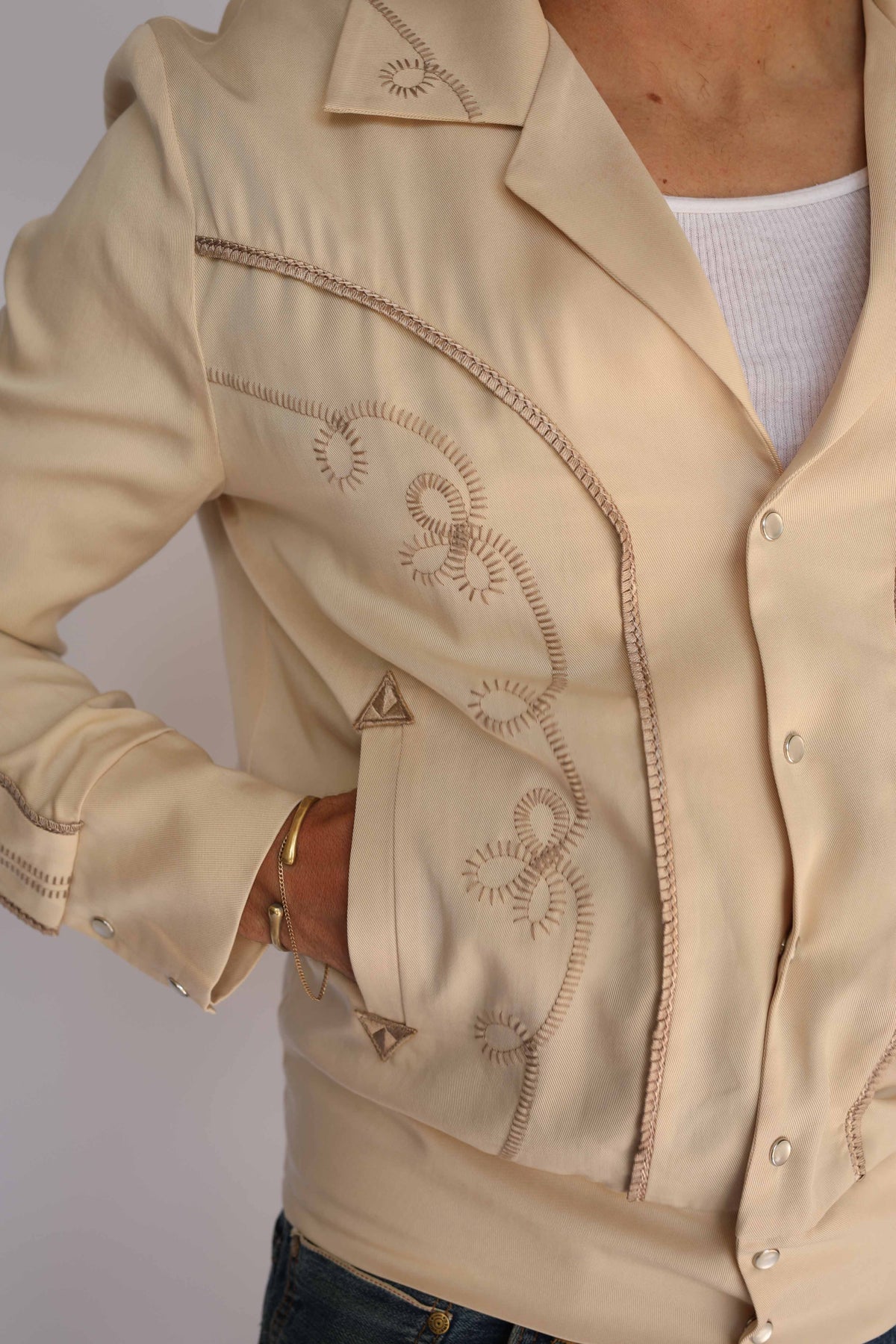A man wearing a cream H Bar C vintage inspired western-style jacket with embroidered details, a white undershirt, and blue jeans, standing against a plain light gray background.