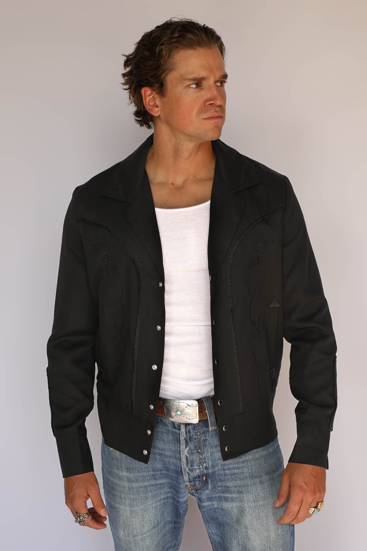 A man wearing a black H Bar C vintage inspired western-style snap-button jacket over a white t-shirt and blue jeans, standing against a plain light gray background.