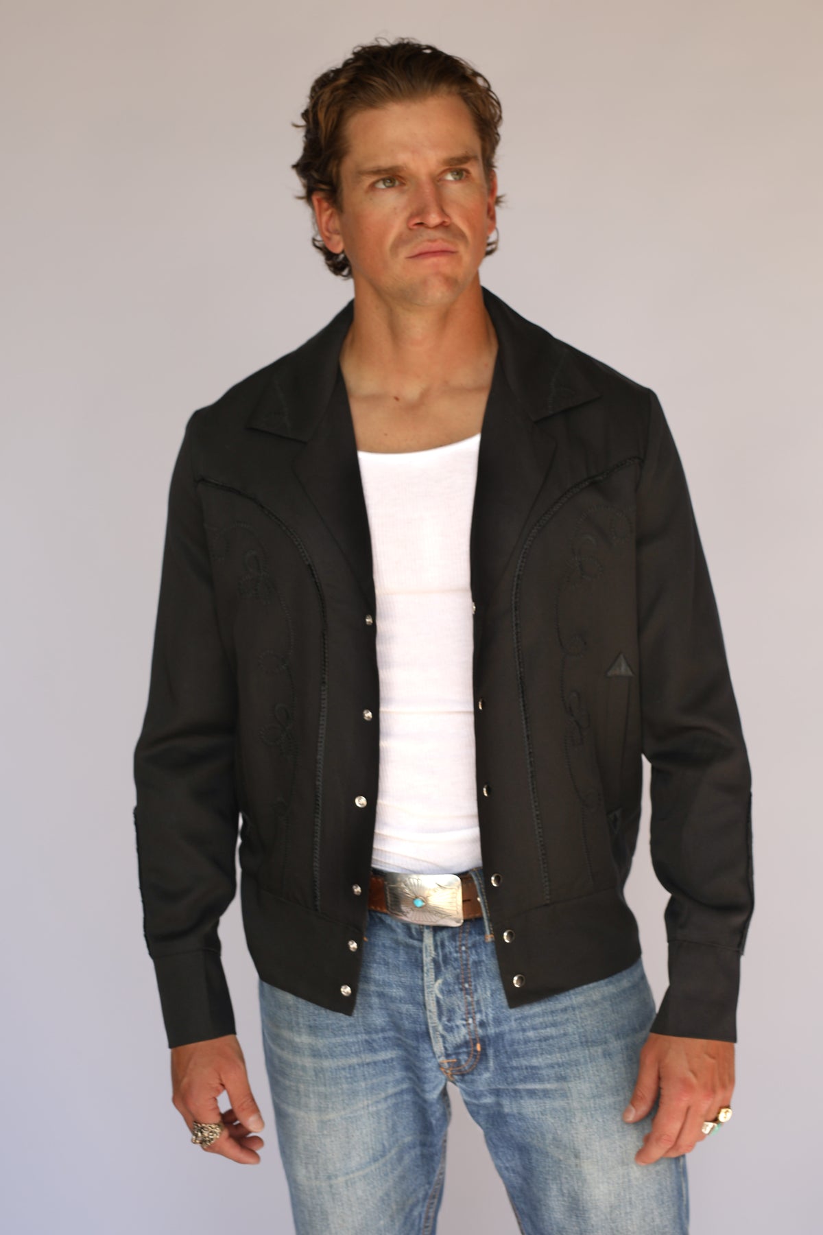 A man wearing a black H Bar C vintage inspired western-style snap-button jacket over a white t-shirt and blue jeans, standing against a plain light gray background.