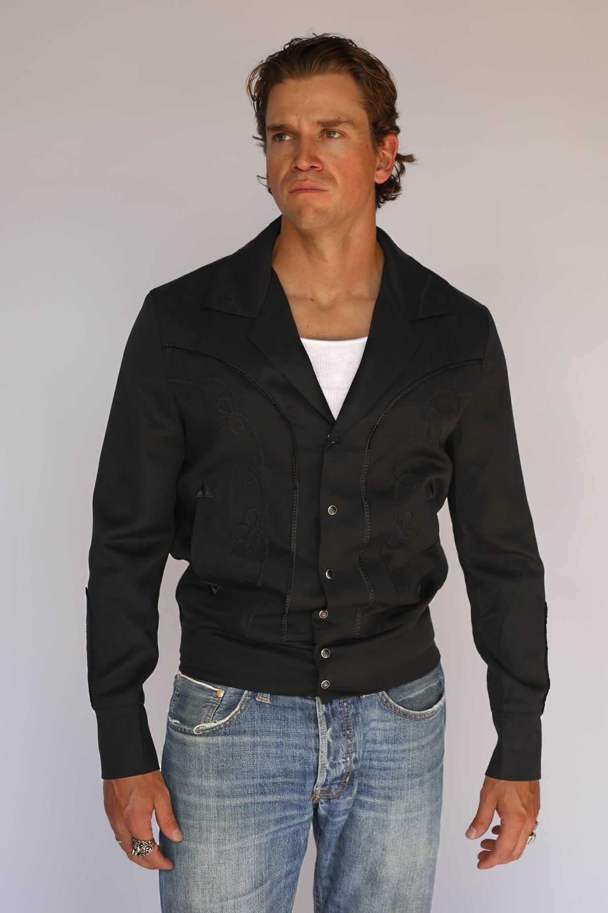 A man wearing a black H Bar C vintage inspired western-style snap-button jacket over a white t-shirt and blue jeans, standing against a plain light gray background.