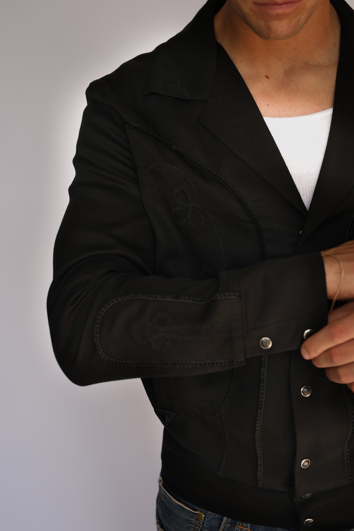 A man wearing a black H Bar C vintage inspired western-style snap-button jacket over a white t-shirt and blue jeans, standing against a plain light gray background.