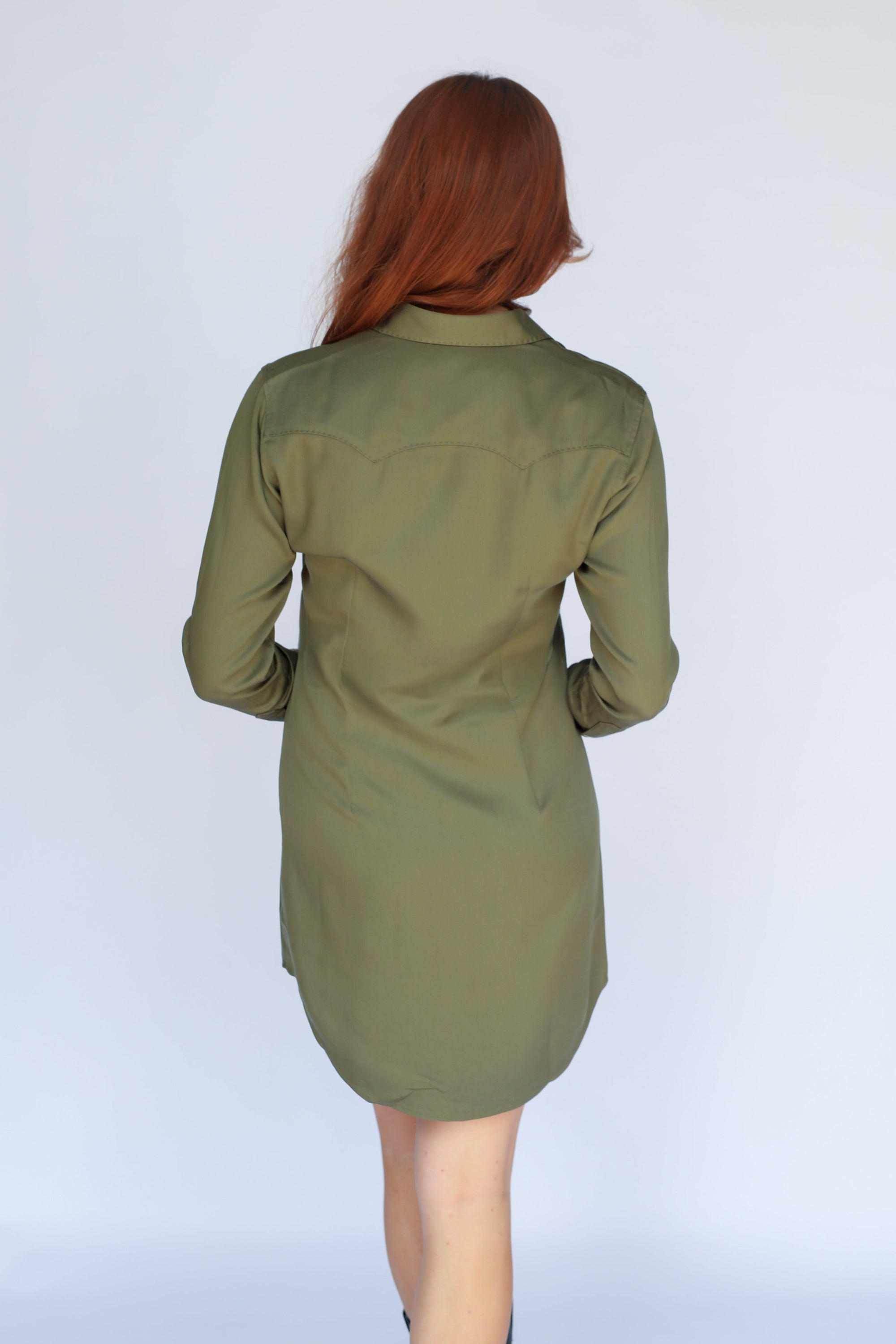 A woman with red hair is shown from the back wearing an olive green long-sleeve shirt dress against a light gray studio background.