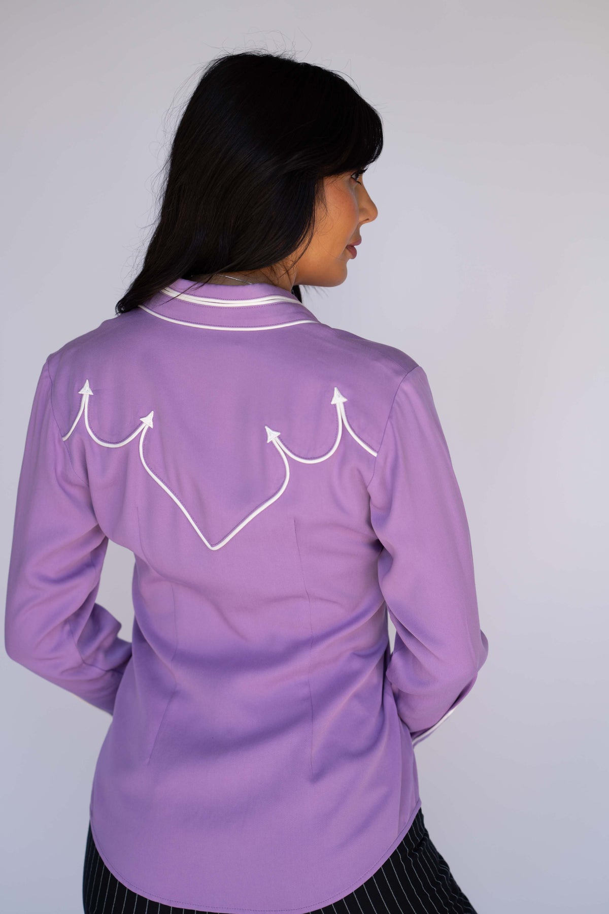 A woman shown from the back wearing a lavender purple long-sleeve shirt with white western-style embroidered arrows on the back, set against a plain light gray background.