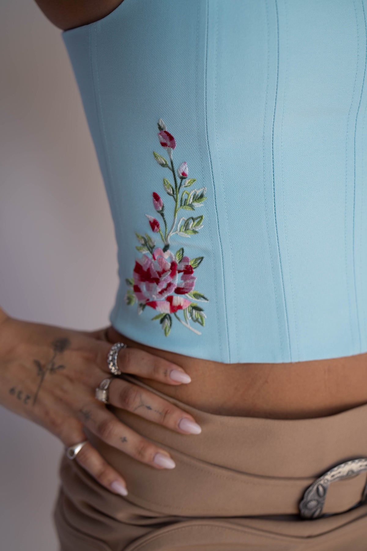 A close-up of a light blue structured corset top with a floral embroidery on the side, worn by a model with a tan skirt and rings.