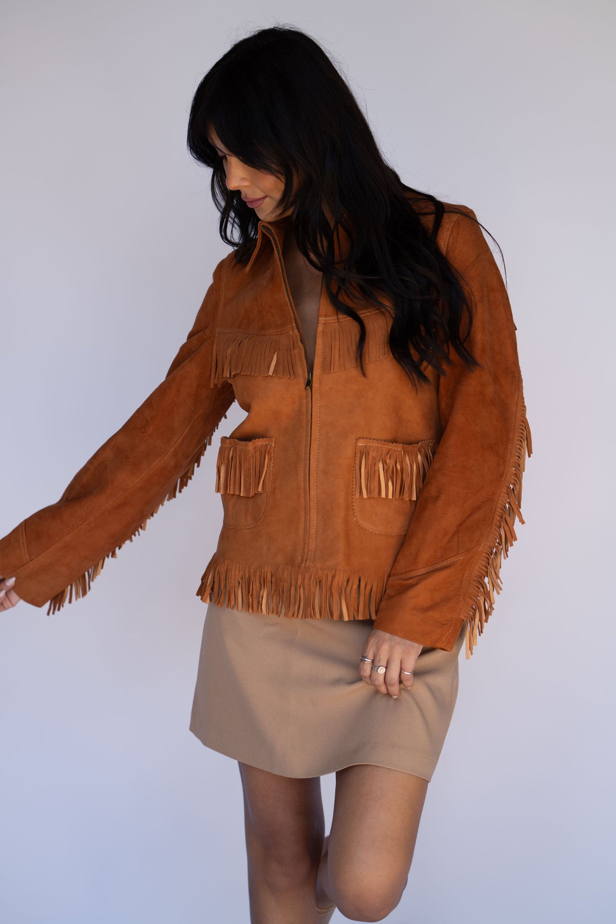 A woman wearing a brown suede fringe jacket and a beige mini skirt, posing against a light gray studio background.