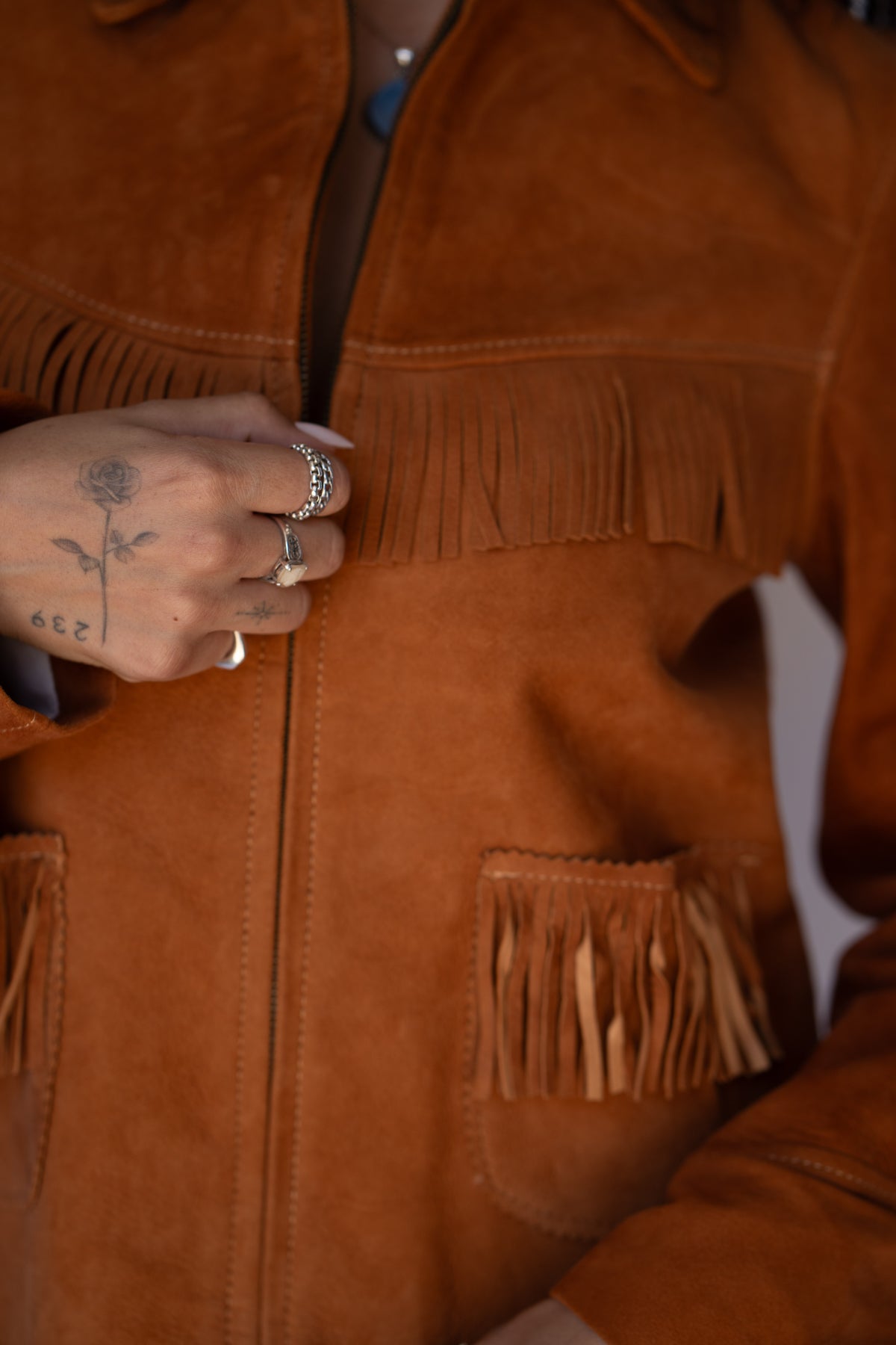 Close-up of a person wearing a warm brown suede fringe jacket, with a tattooed hand adjusting the front zipper and wearing silver rings.