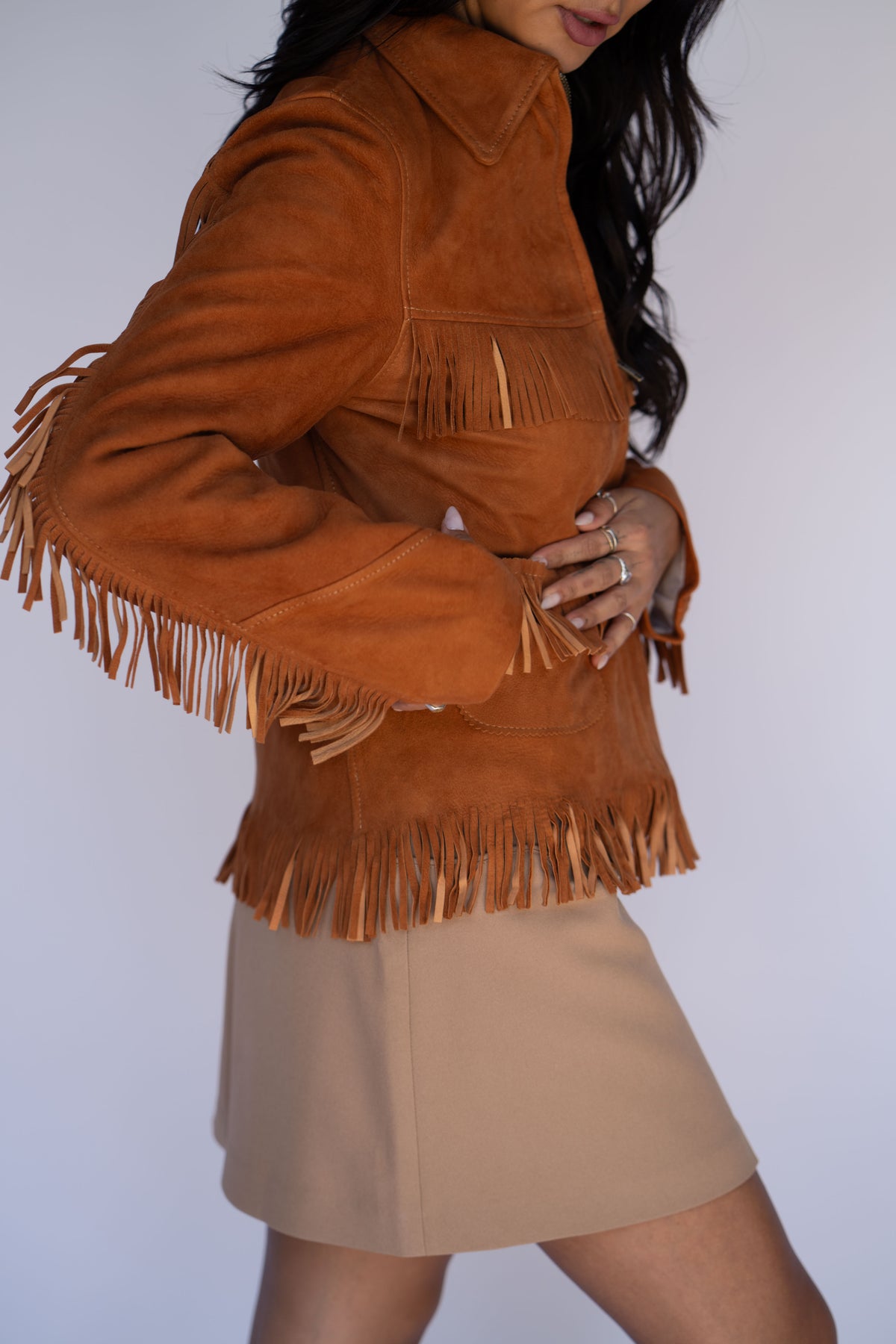 A woman wearing a cropped tan suede fringe jacket with a high collar and a beige mini skirt, photographed against a plain light background.