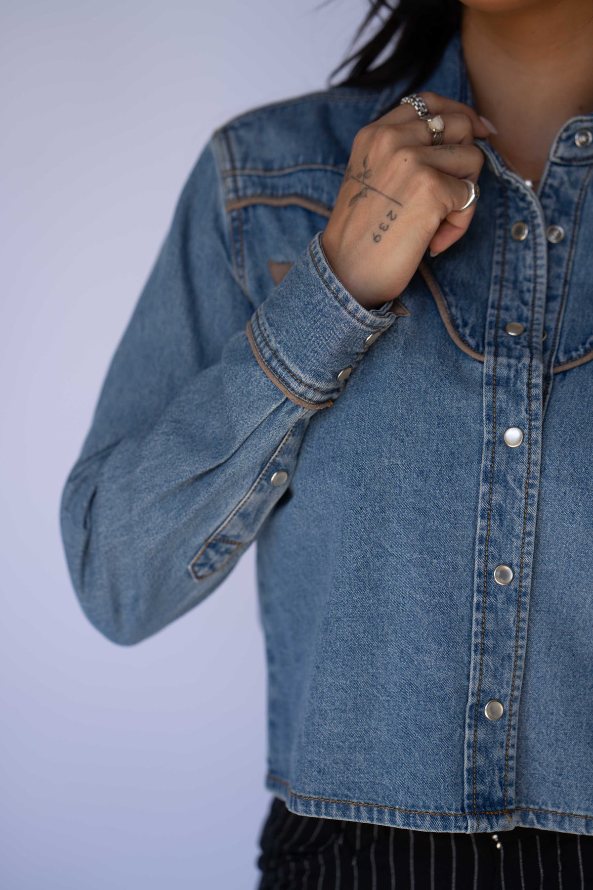 A close-up of a person wearing a cropped blue denim jacket with silver buttons, featuring a western-style chest pocket and a buttoned cuff, set against a solid light gray background.
