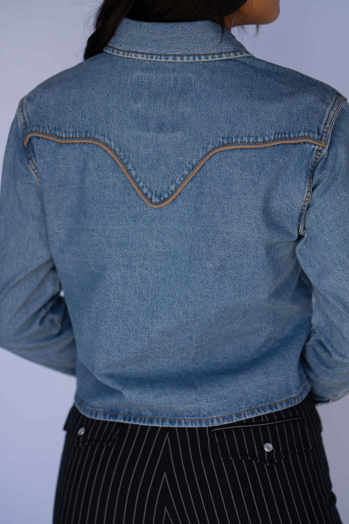 A person shown from the back wearing a cropped light blue denim jacket with contrast tan stitching and dark pinstriped pants against a plain light background.