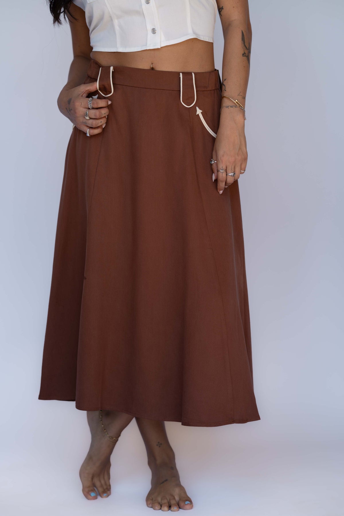 A woman wearing a brown midi skirt with white drawstring details, paired with a white cropped top, standing against a plain light background.