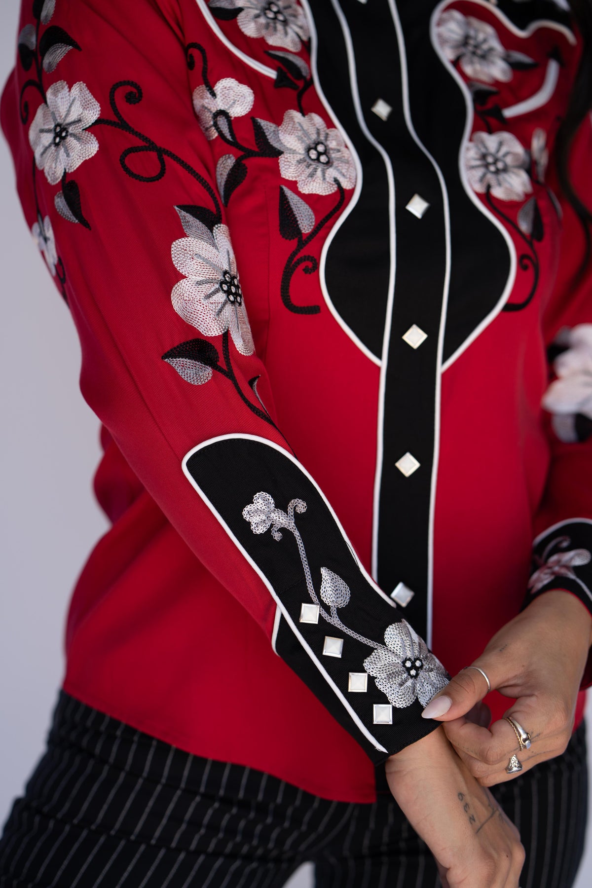 A close-up of a person wearing a bright red blouse with black and white floral embroidery and mirror-like diamond accents on the front placket and sleeve cuffs, paired with black-and-white striped pants.