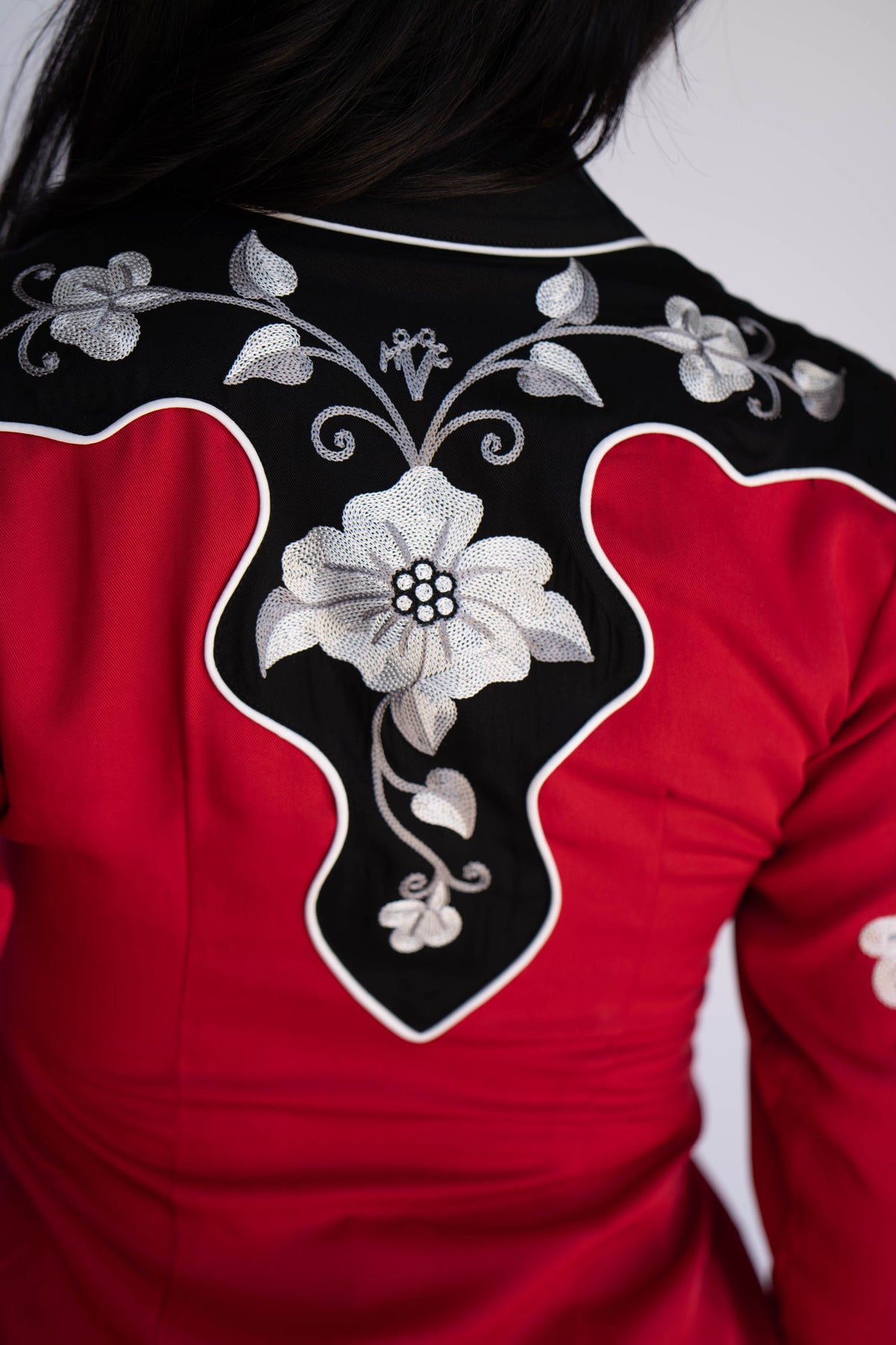 Close-up of the back of a red and black embroidered garment, showing a large white floral motif with silver-gray detailing on a black panel outlined in white, worn by a person.