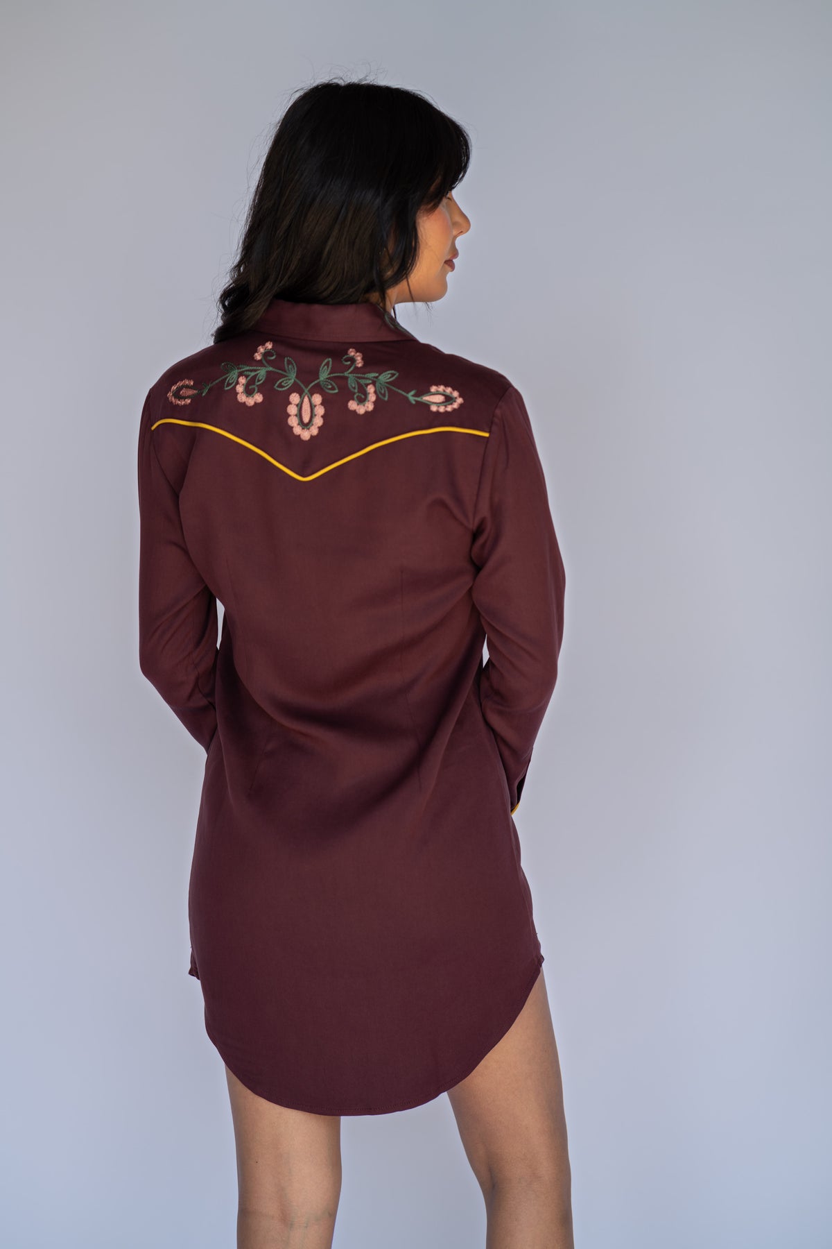 A woman shown from the back wearing a long-sleeve burgundy western-style shirt dress with embroidered details on the shoulders, set against a plain light gray background.