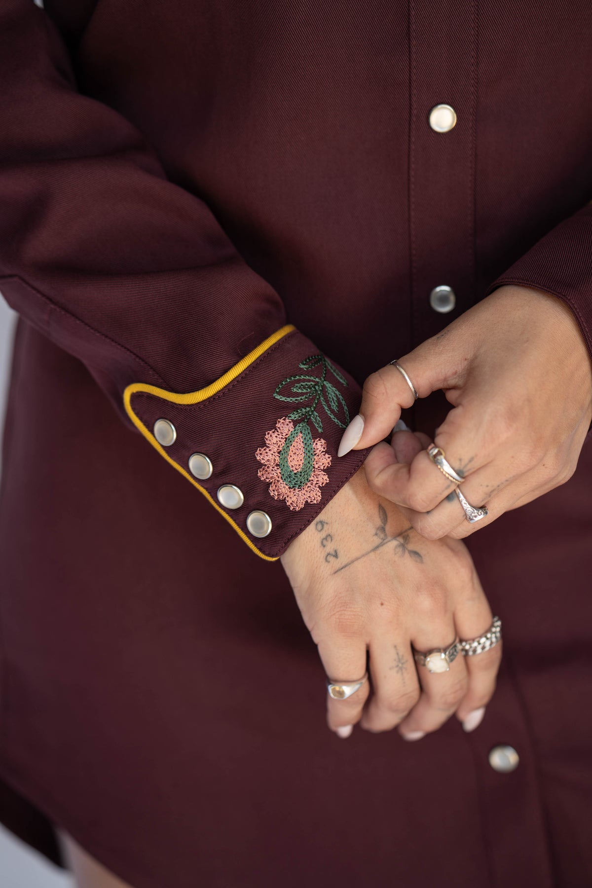 Close-up of a person wearing a burgundy button-front top or dress, focusing on the embroidered and buttoned cuff. The cuff has a floral embroidery in pink, green, and white with yellow piping, and several silver-tone buttons are visible.
