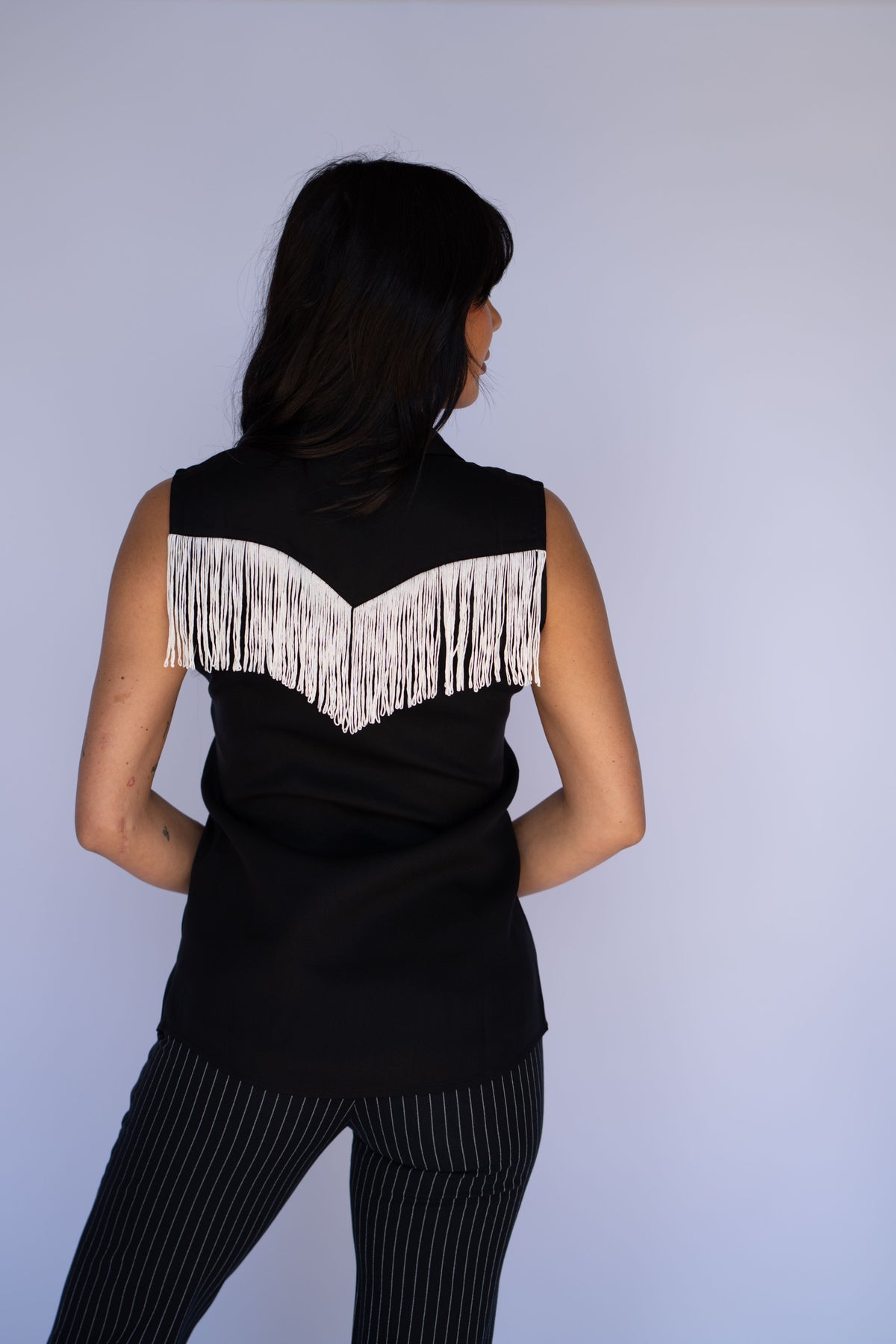 A woman shown from the back wearing a sleeveless black top with a wide white fringe detail across the shoulders and black pinstriped pants, set against a plain light gray studio background.