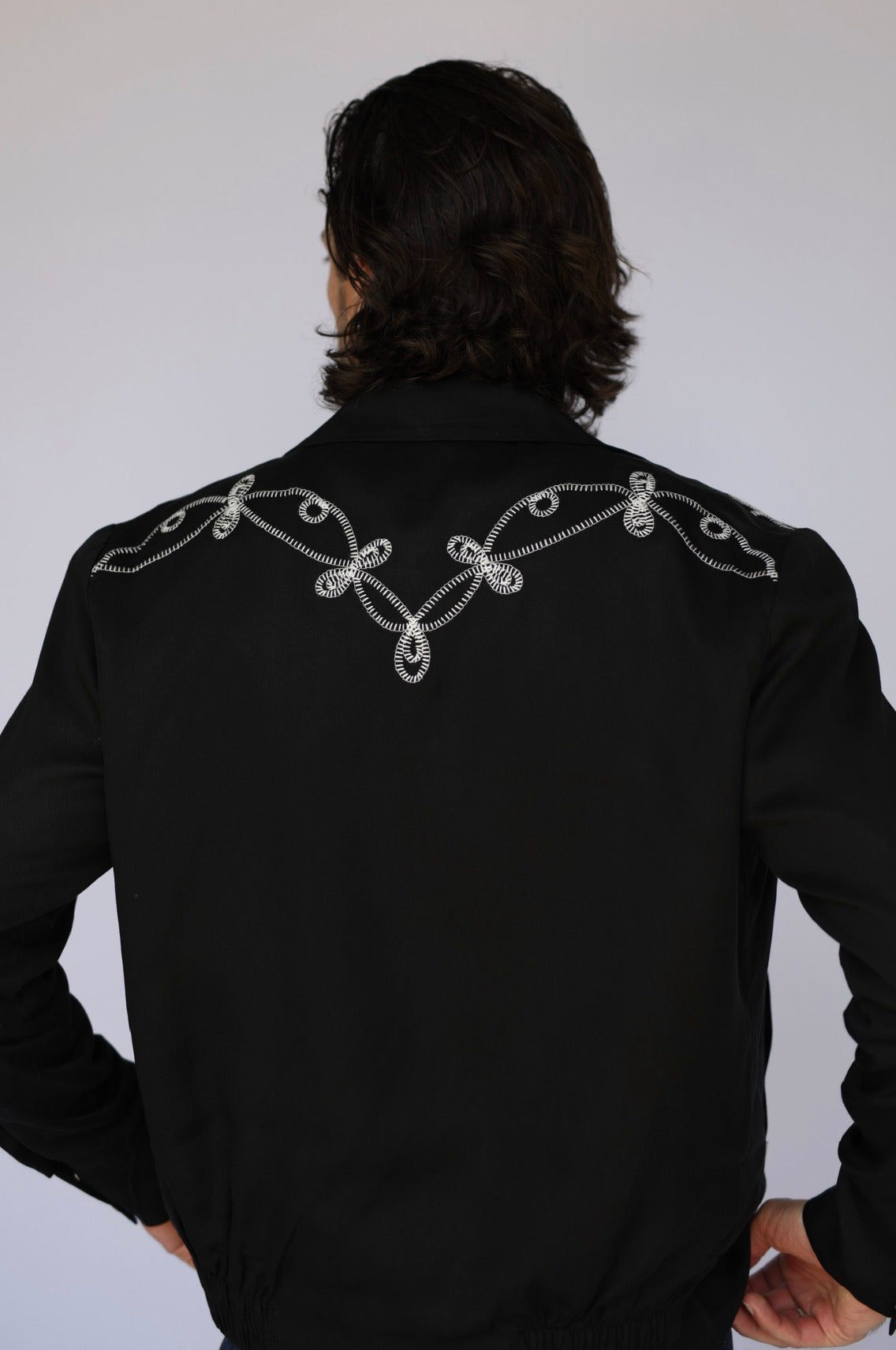 A man viewed from behind wearing a black western-style jacket with intricate silver embroidered scrollwork across the shoulders, set against a plain light gray background.