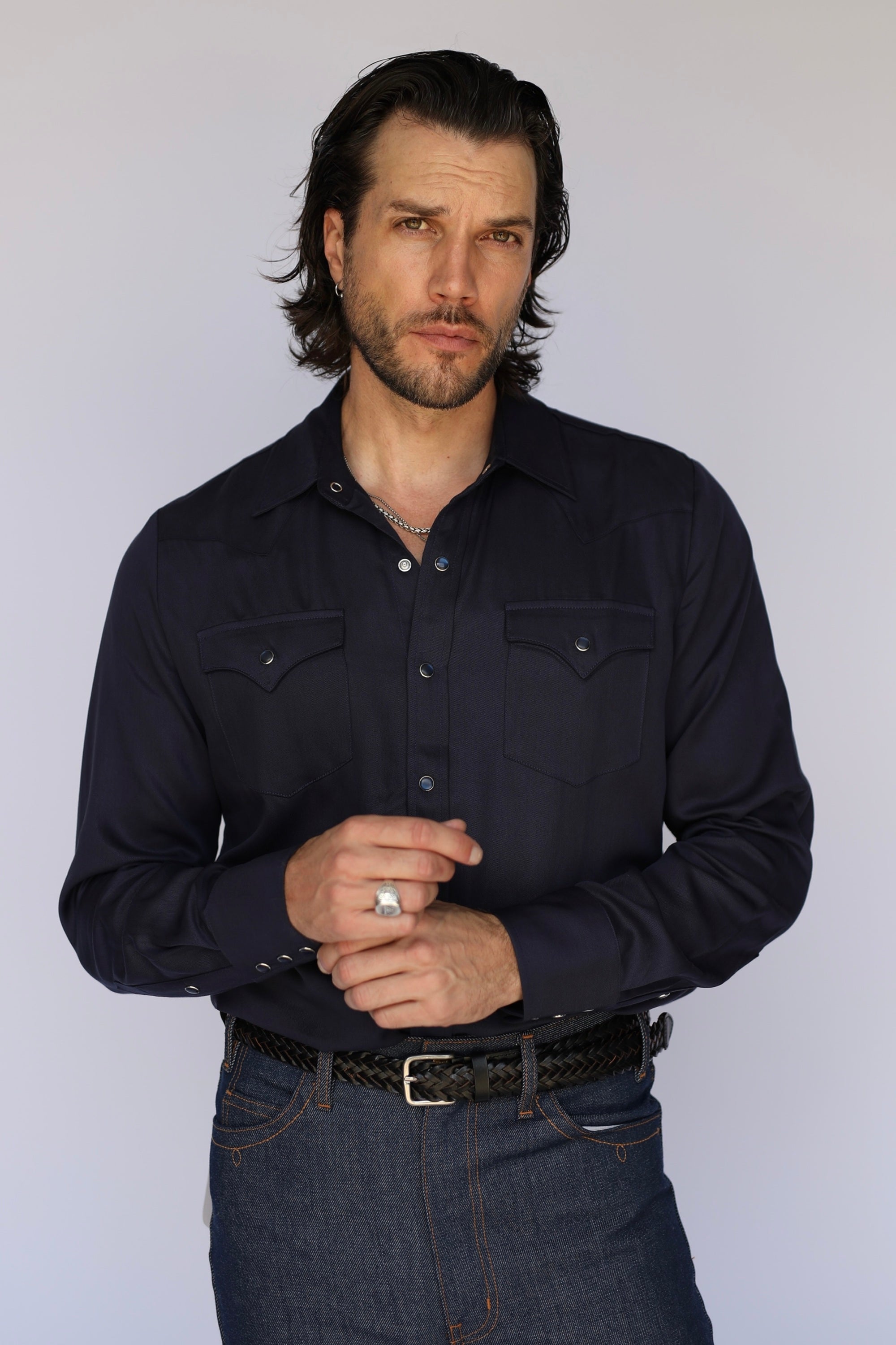 A man wearing a dark navy long-sleeve western-style button-down shirt with chest pockets, paired with dark blue jeans and a black braided belt, standing against a light gray studio background.