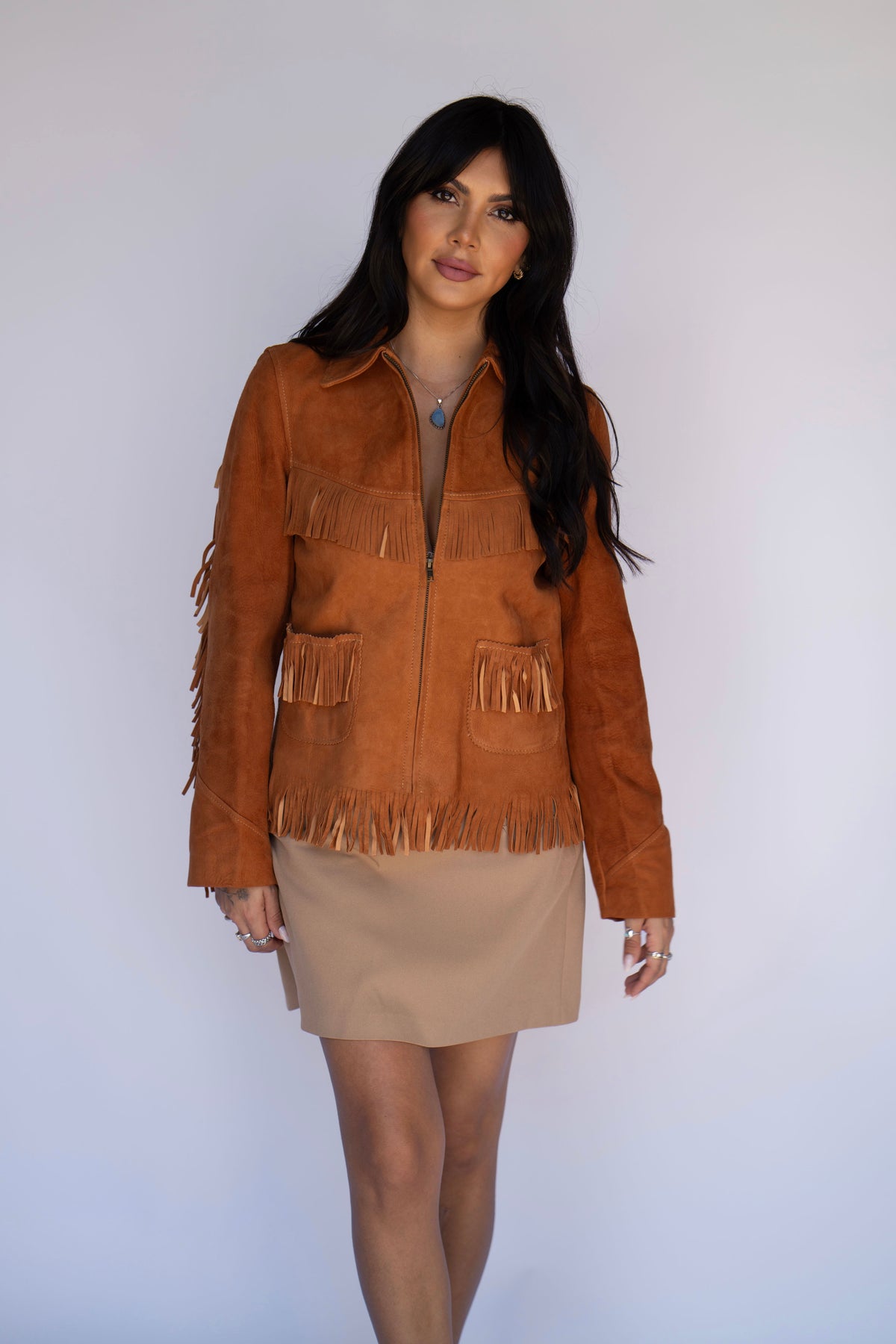 A woman modeling a brown suede fringe jacket over a beige mini skirt, standing against a plain light gray background.