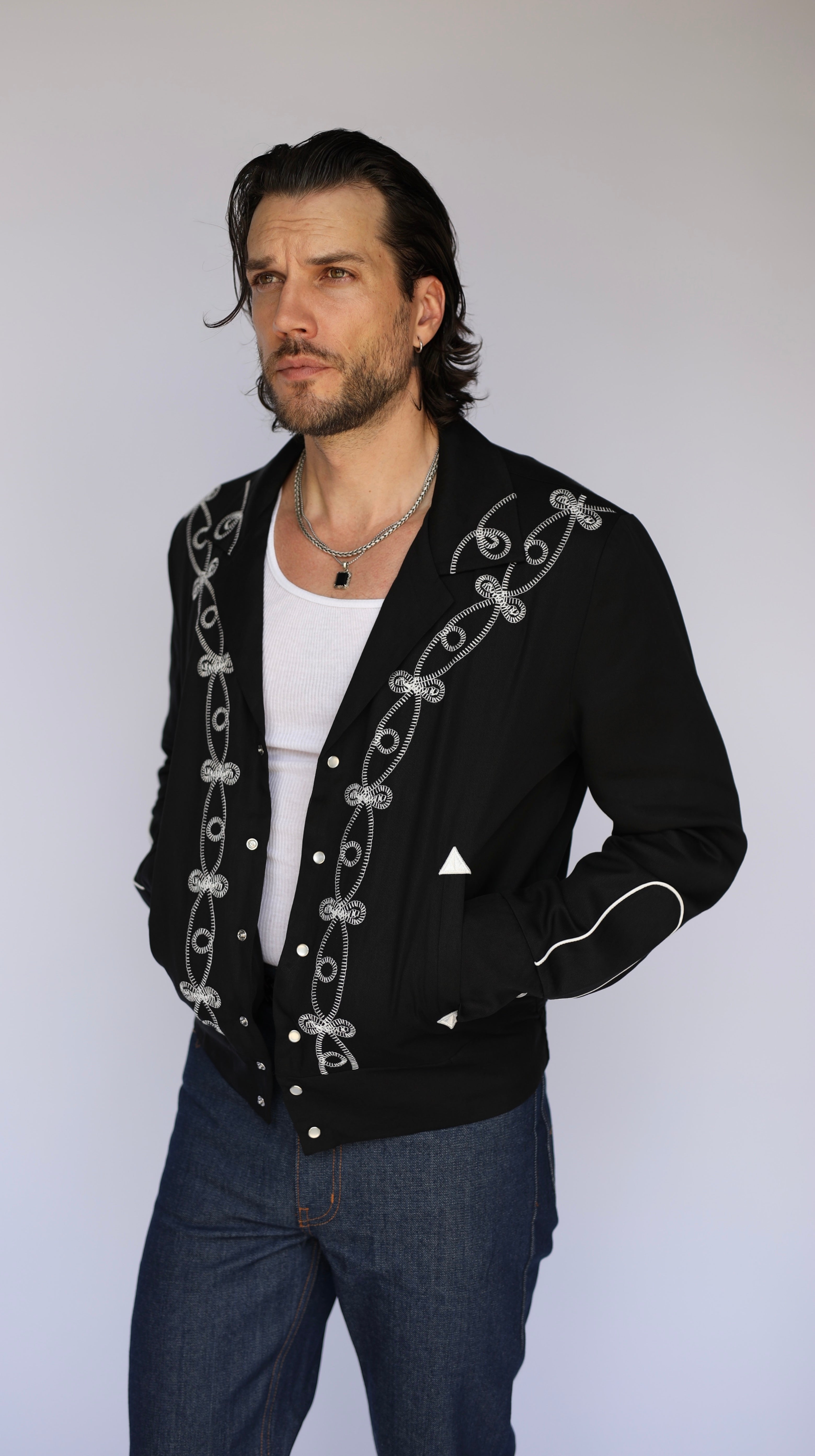 A man wearing a black western-style cardigan with white embroidered scrollwork and snap buttons, layered over a white t-shirt and blue jeans, photographed against a light gray studio background.