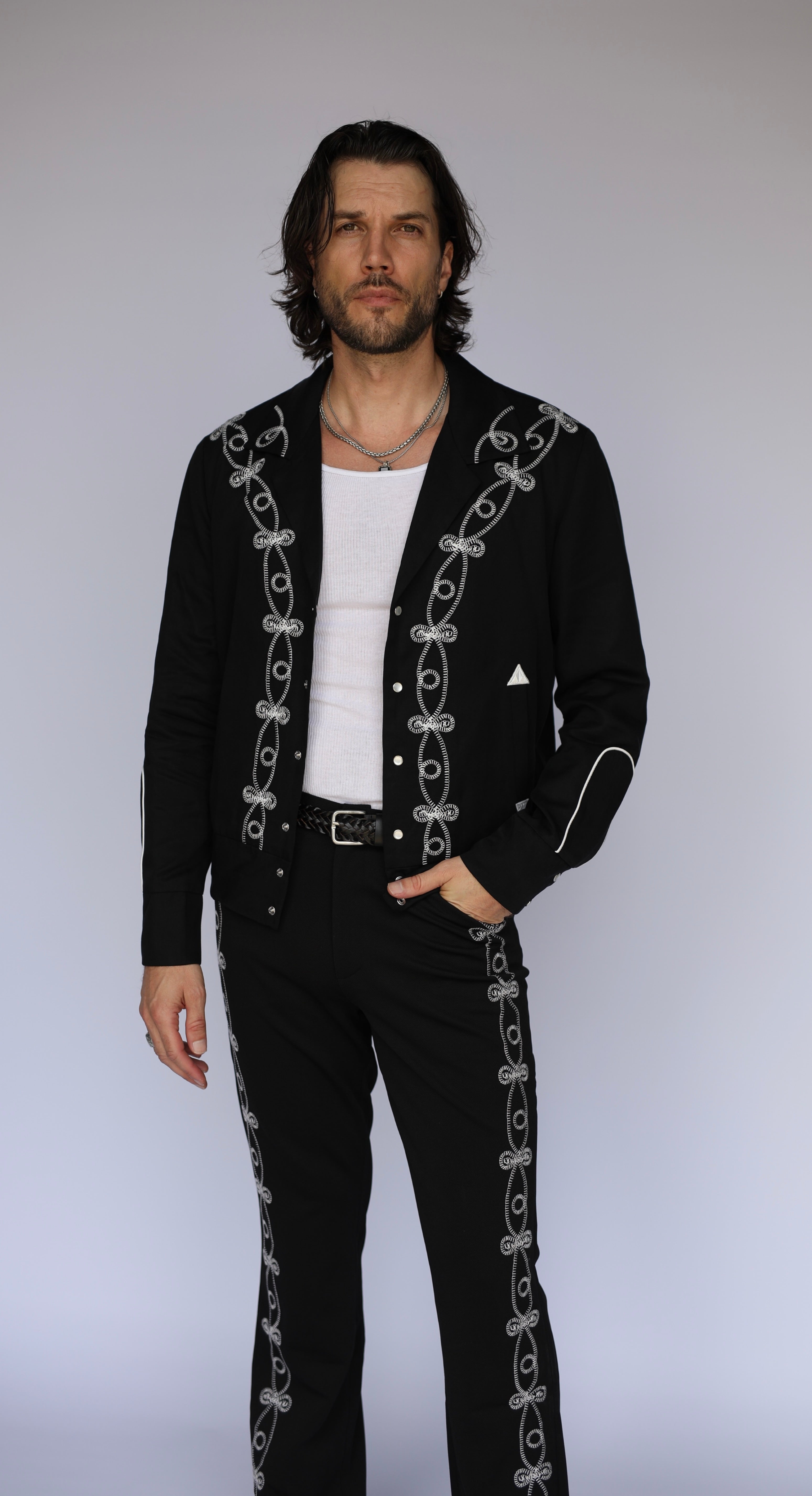 A man modeling a black western-style cardigan and matching pants set, both featuring white embroidered decorative patterns down the front and sides, styled over a white t-shirt and black trousers.