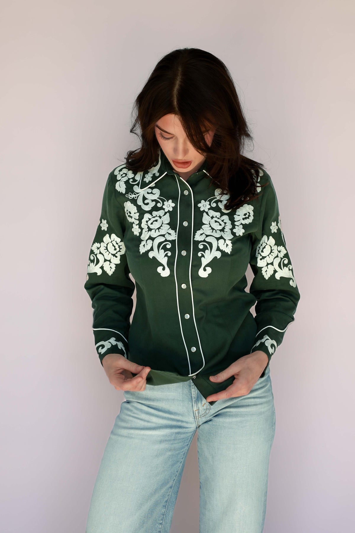 A woman wearing a dark green long-sleeve button-up shirt with white floral embroidery on the chest, shoulders, and cuffs, paired with light-wash blue jeans, set against a plain light gray background.