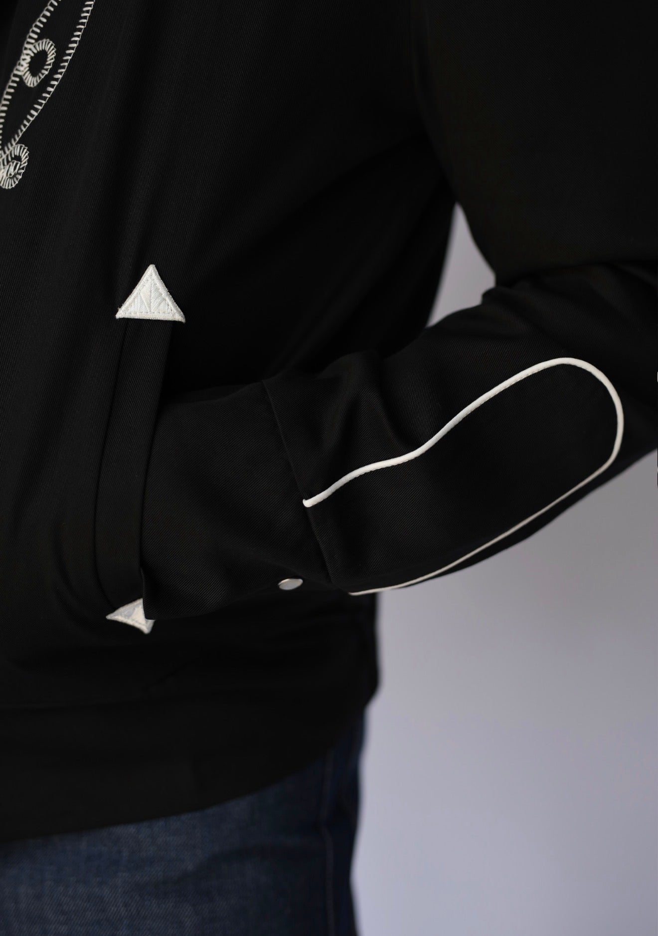 Close-up of a black jacket with white contrast piping and white geometric trim details, worn by a model.