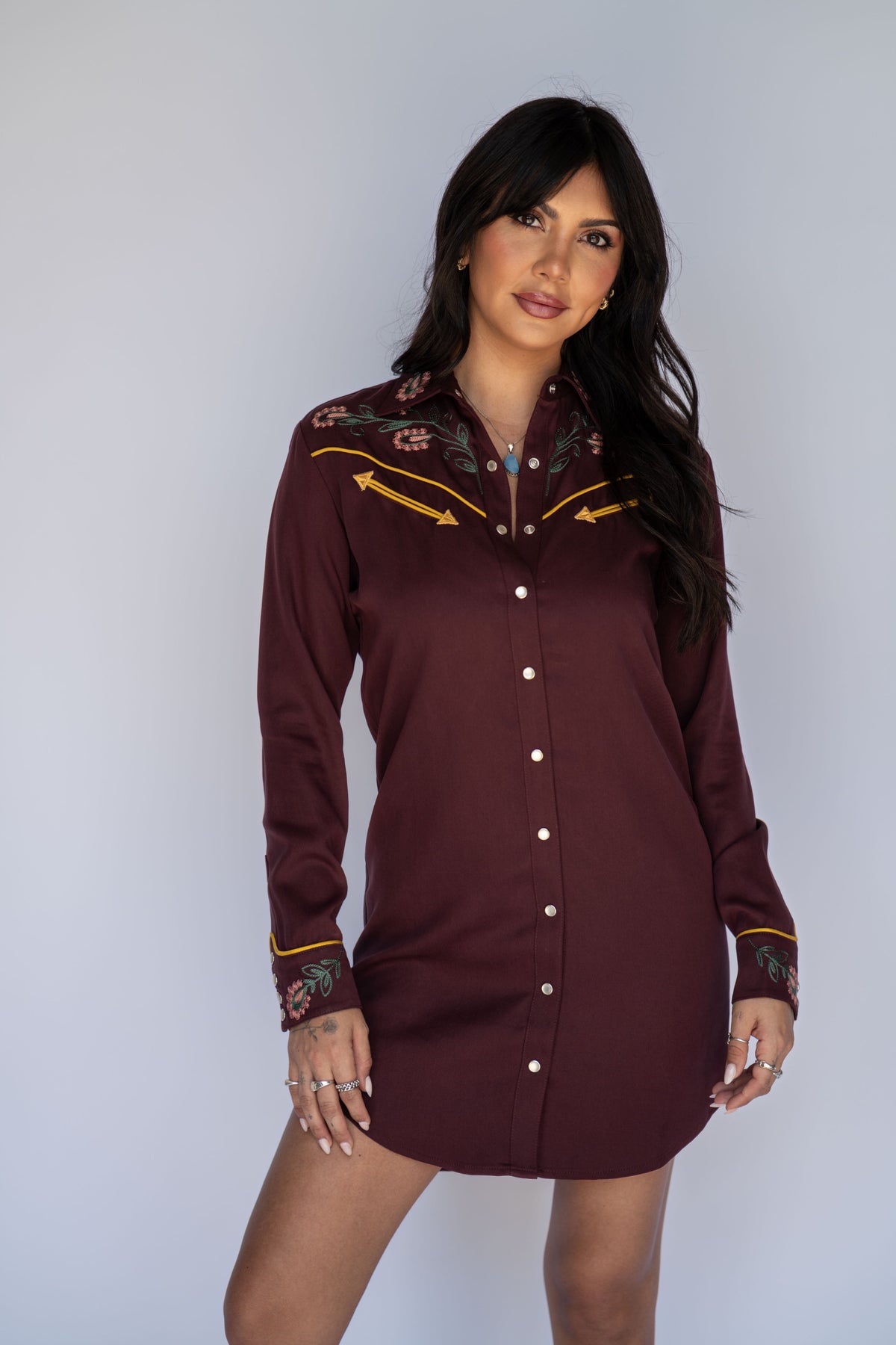A woman modeling a long-sleeve western-style mini dress in deep burgundy. The dress features a button-down front, embroidered floral details on the chest and cuffs, and yellow embroidered accents on the shoulders, set against a plain light gray background.