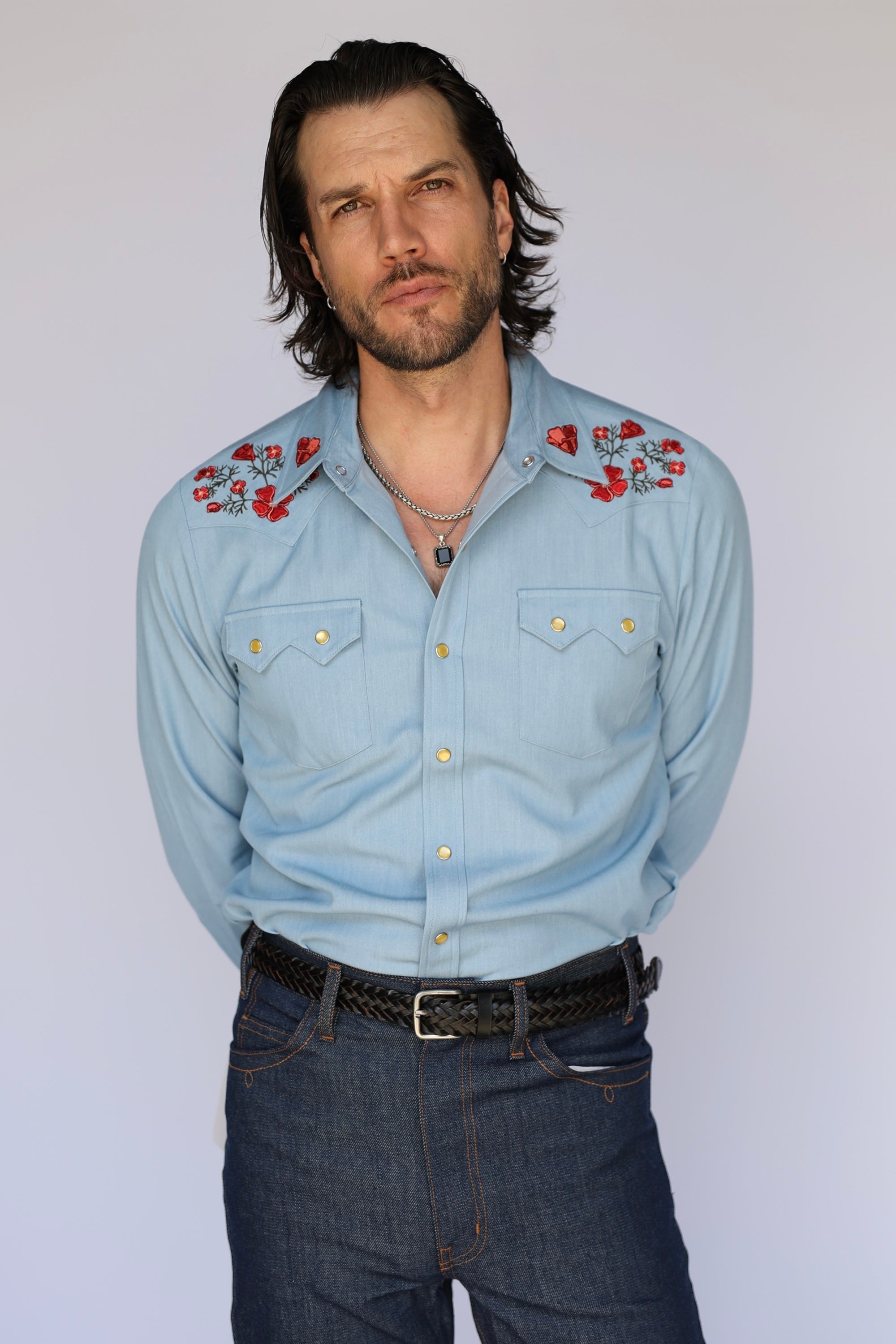 A man wearing a light blue western-style button-up shirt with red and white floral embroidery on the shoulders, paired with dark blue jeans and a black braided belt, standing against a plain light gray background.
