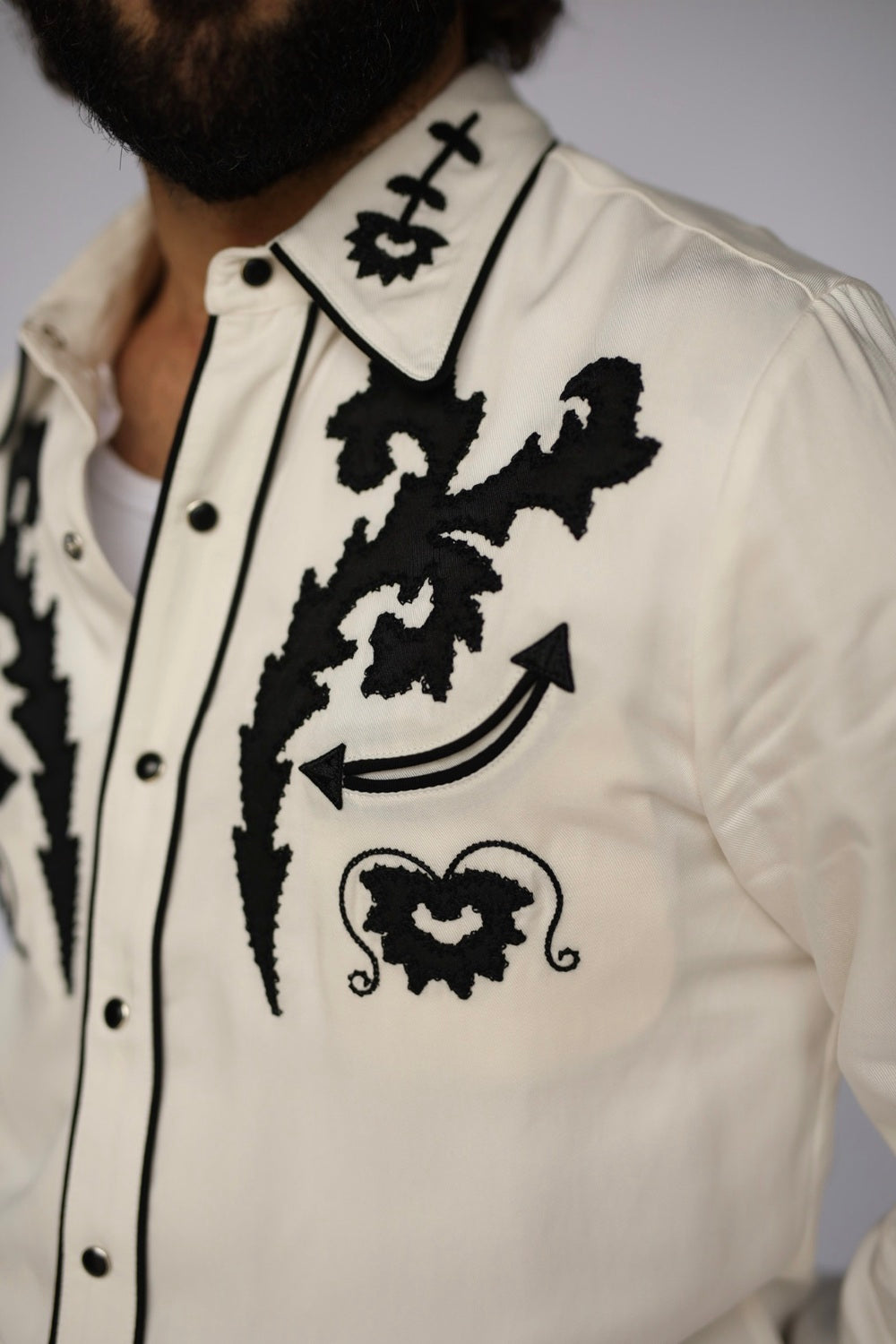 Close-up of a man wearing a cream western-style button-up shirt with bold black embroidered floral and arrow motifs on the chest, plus black piping along the collar and placket.