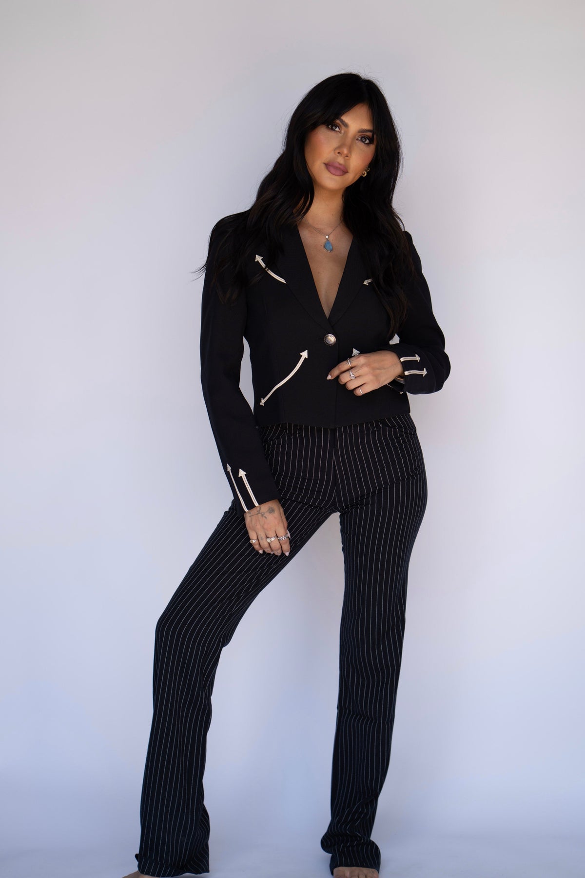 A woman modeling a black two-piece outfit consisting of a long-sleeve cropped blazer with silver zipper details and matching black pinstriped trousers, posed against a plain light gray studio background.