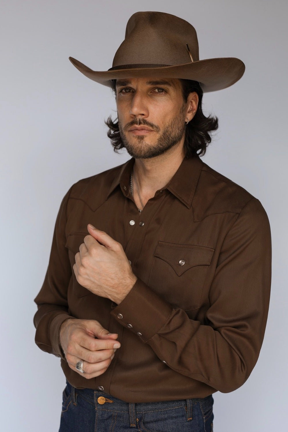 A man wearing a brown long-sleeve western-style button-up shirt and a matching brown cowboy hat, posed against a light gray studio background.