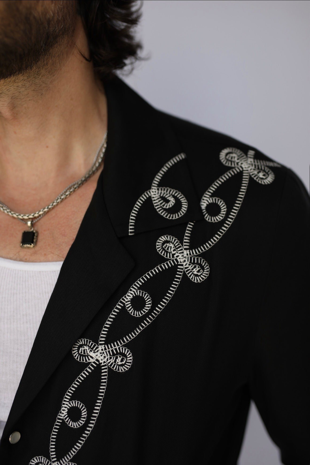 Close-up of a person wearing a black jacket with intricate white embroidered swirl patterns down the shoulder and chest, layered over a white shirt and a silver chain necklace.