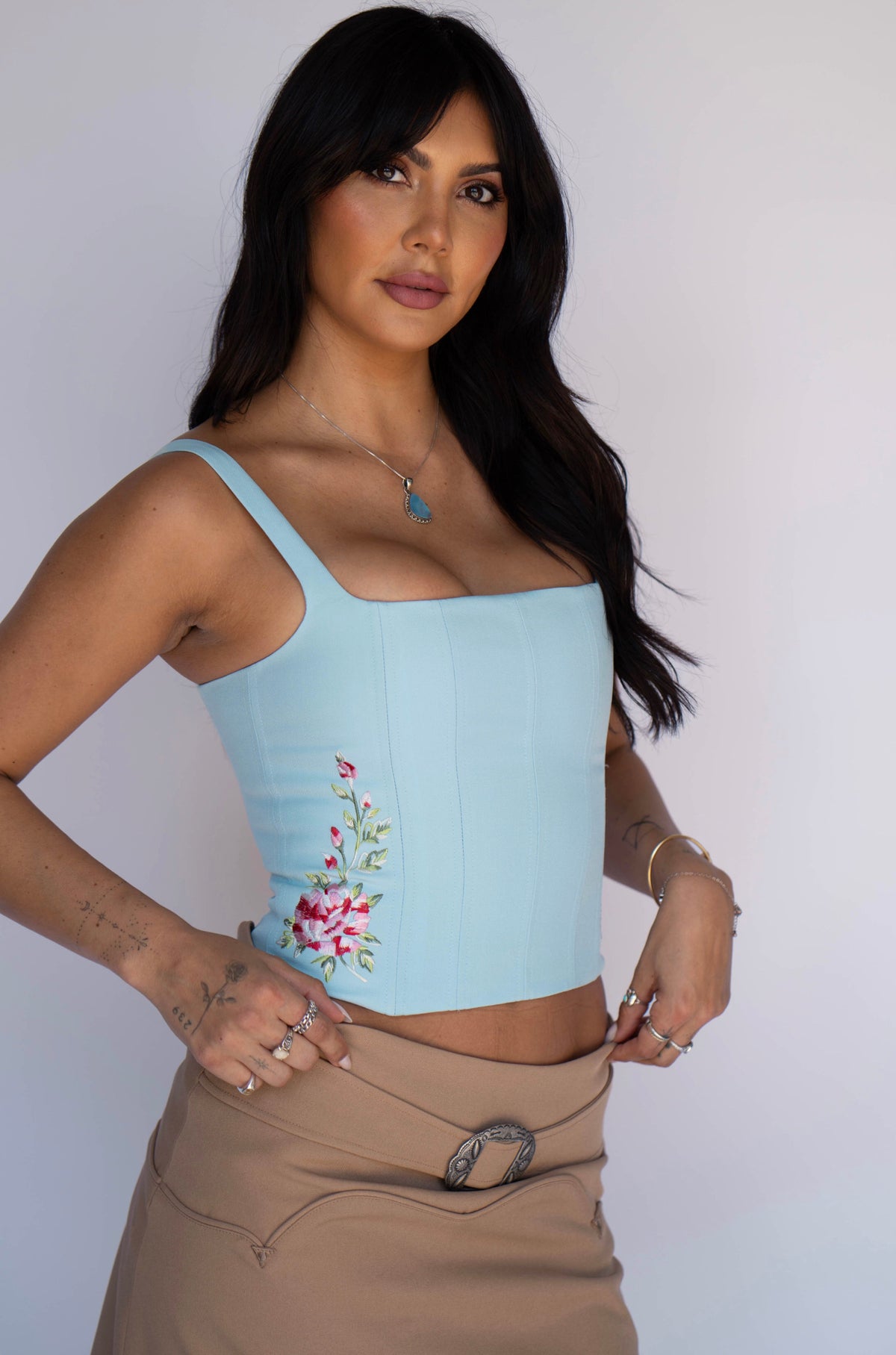 A woman modeling a light blue corset-style camisole top with floral embroidery on the lower left side, paired with a tan skirt, against a plain light gray background.