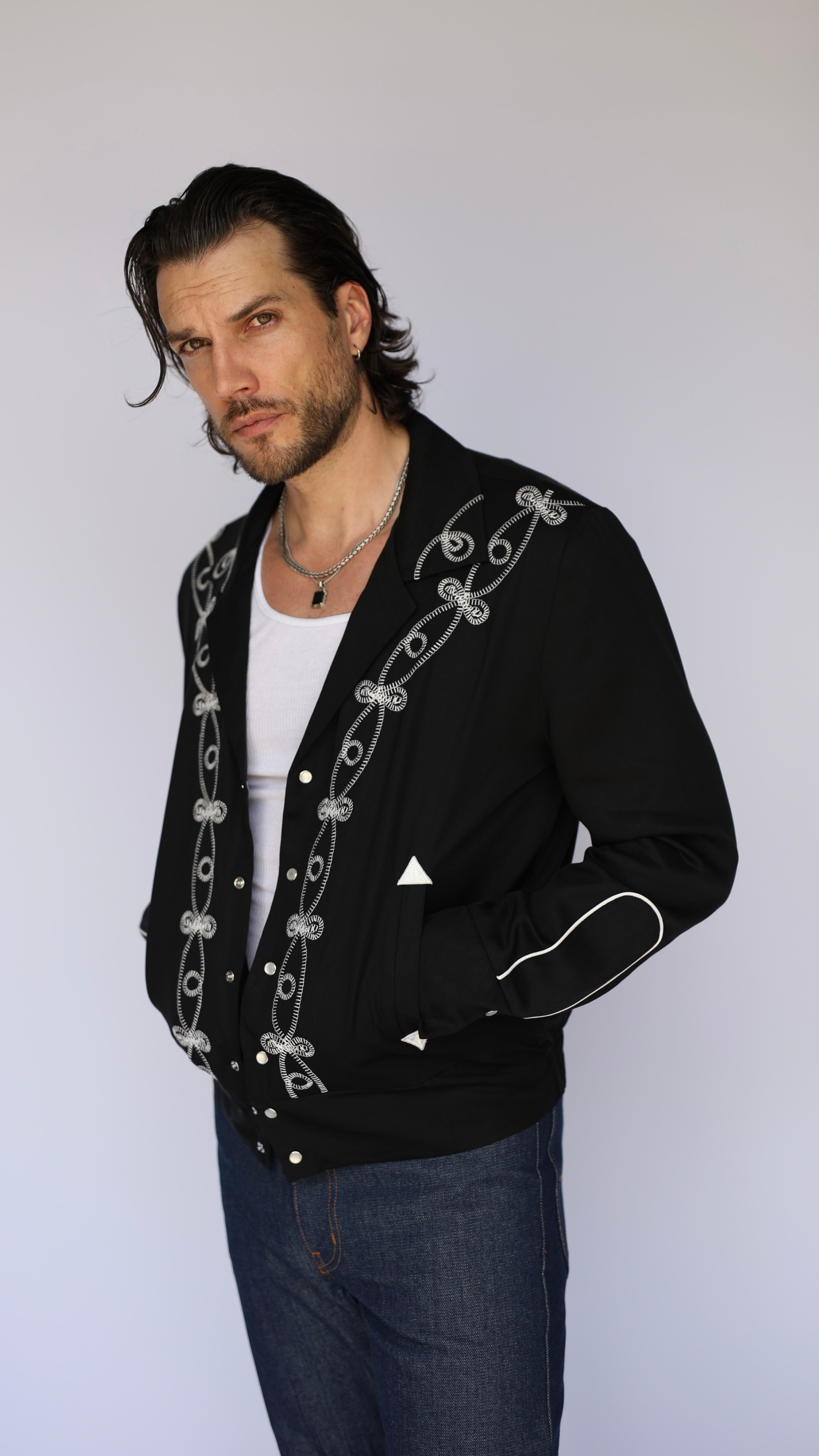 A man wearing a black western-style cardigan jacket with white embroidered details, a white t-shirt, and blue jeans, photographed against a light gray studio background.