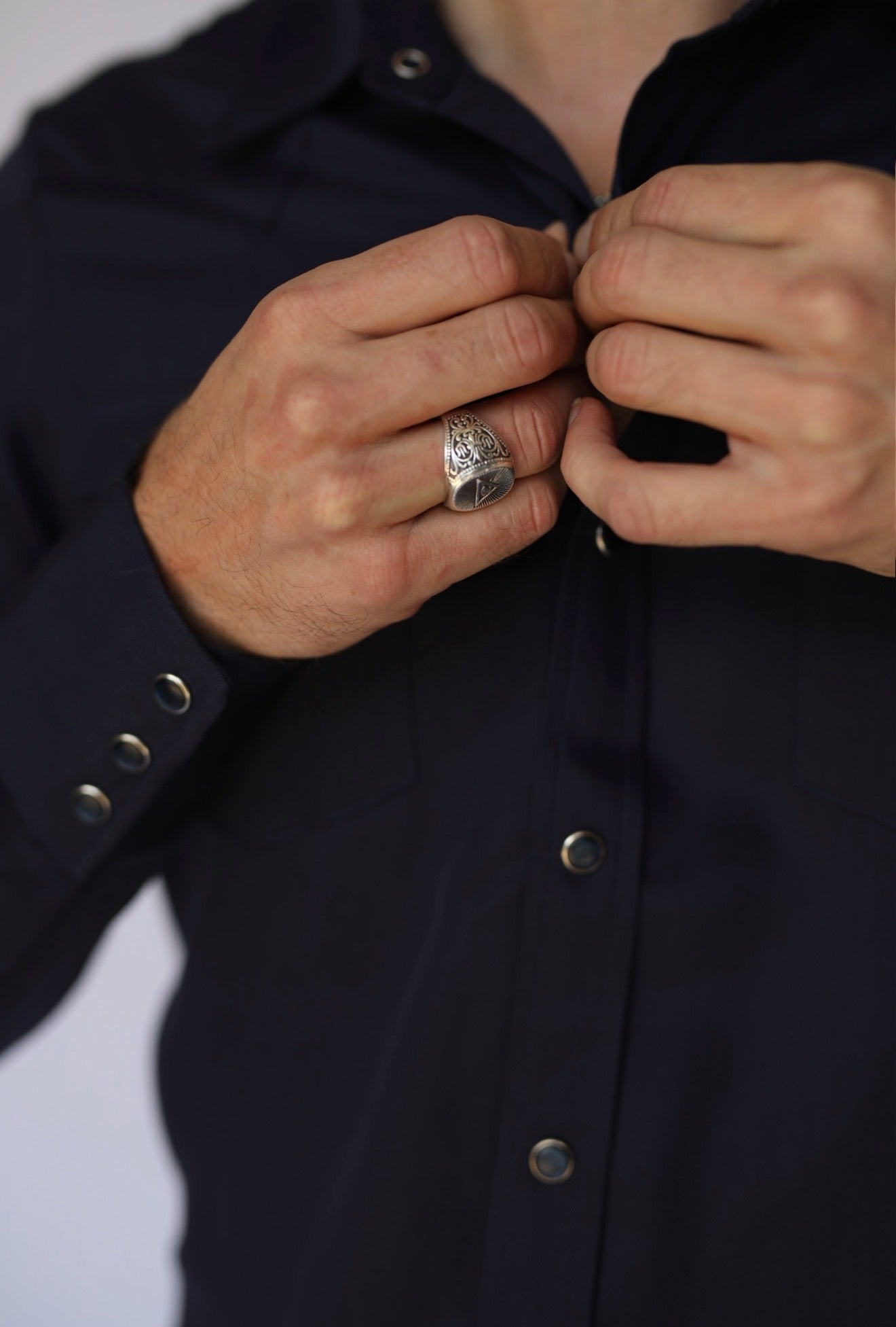 A close-up of a person's hands buttoning a dark navy blue shirt, with a detailed silver ring worn on one finger.