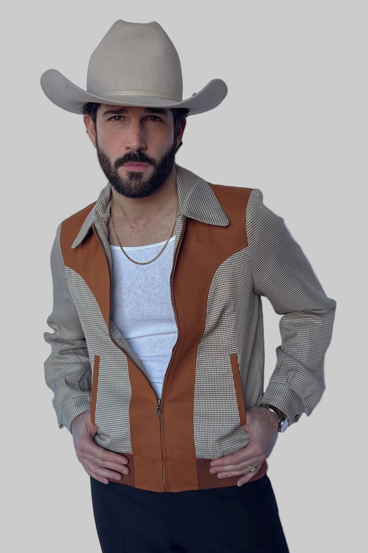 A man wearing a beige cowboy hat and a western-style jacket with tan and gray striped panels over a white t-shirt and dark pants, posed against a solid gray background.