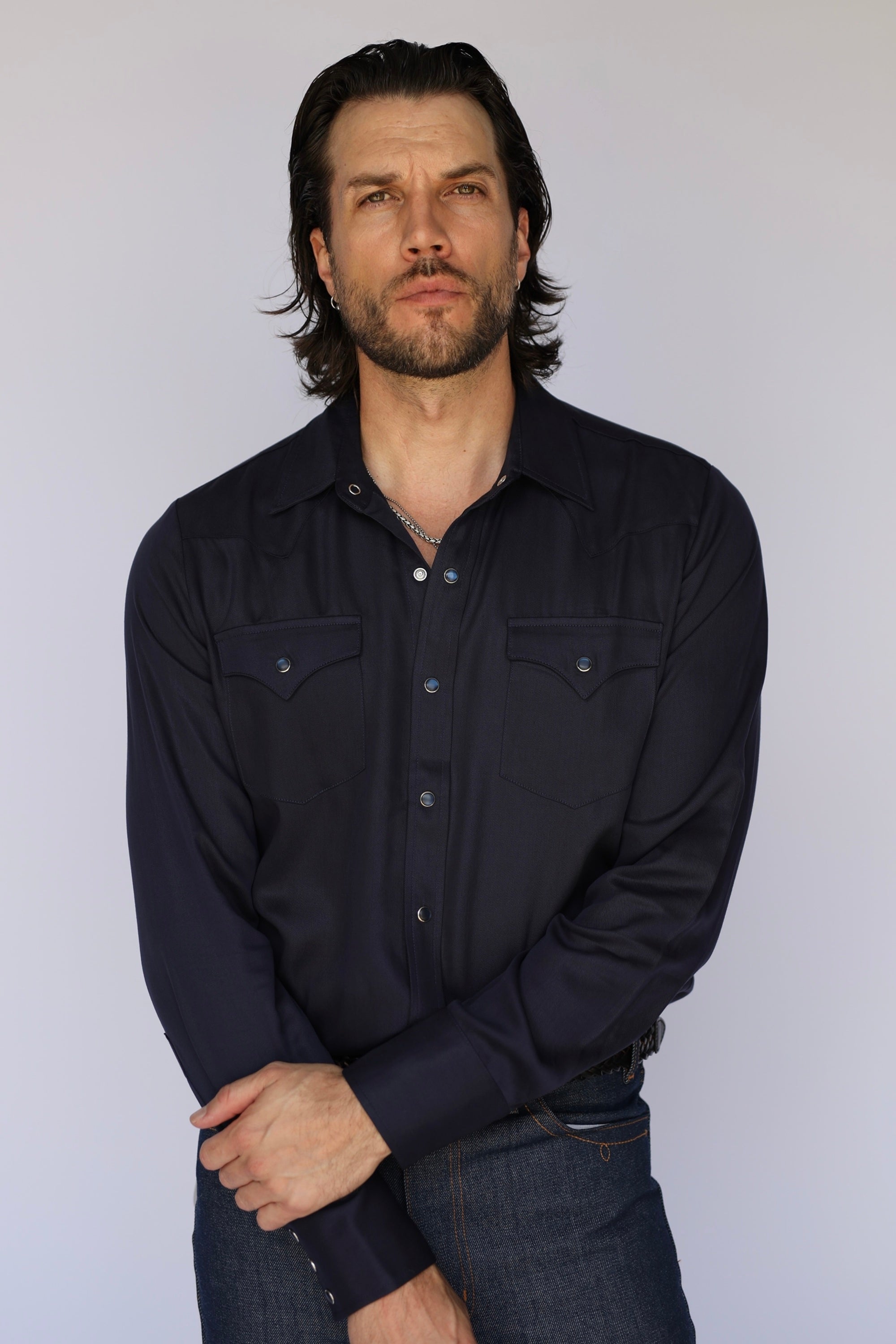 A man wearing a dark navy long-sleeve western-style button-up shirt and dark jeans, standing against a plain light gray background.