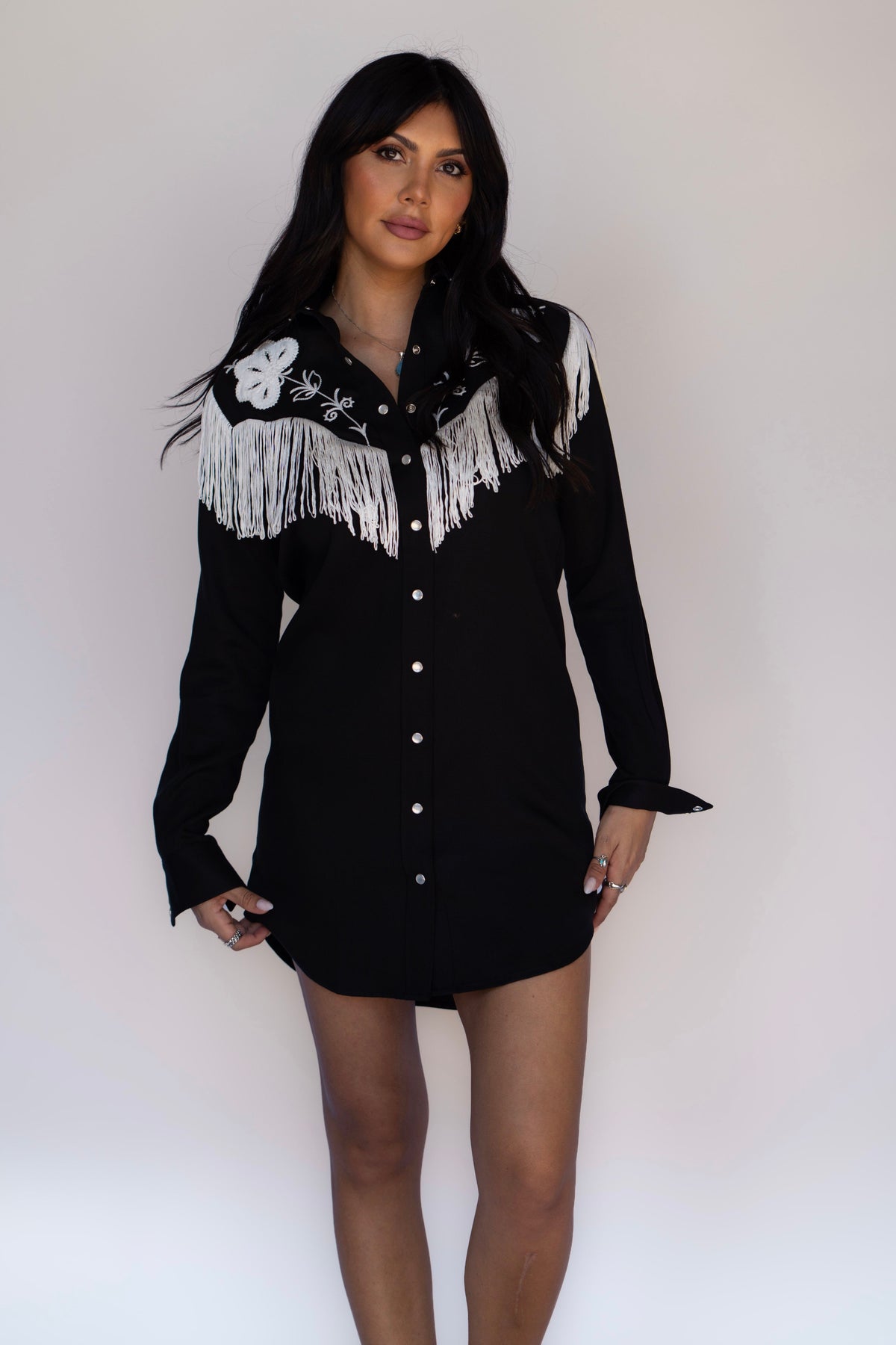 A woman modeling a black long-sleeve western-style shirt dress with white fringe and embroidery on the chest, posed against a plain light gray studio background.