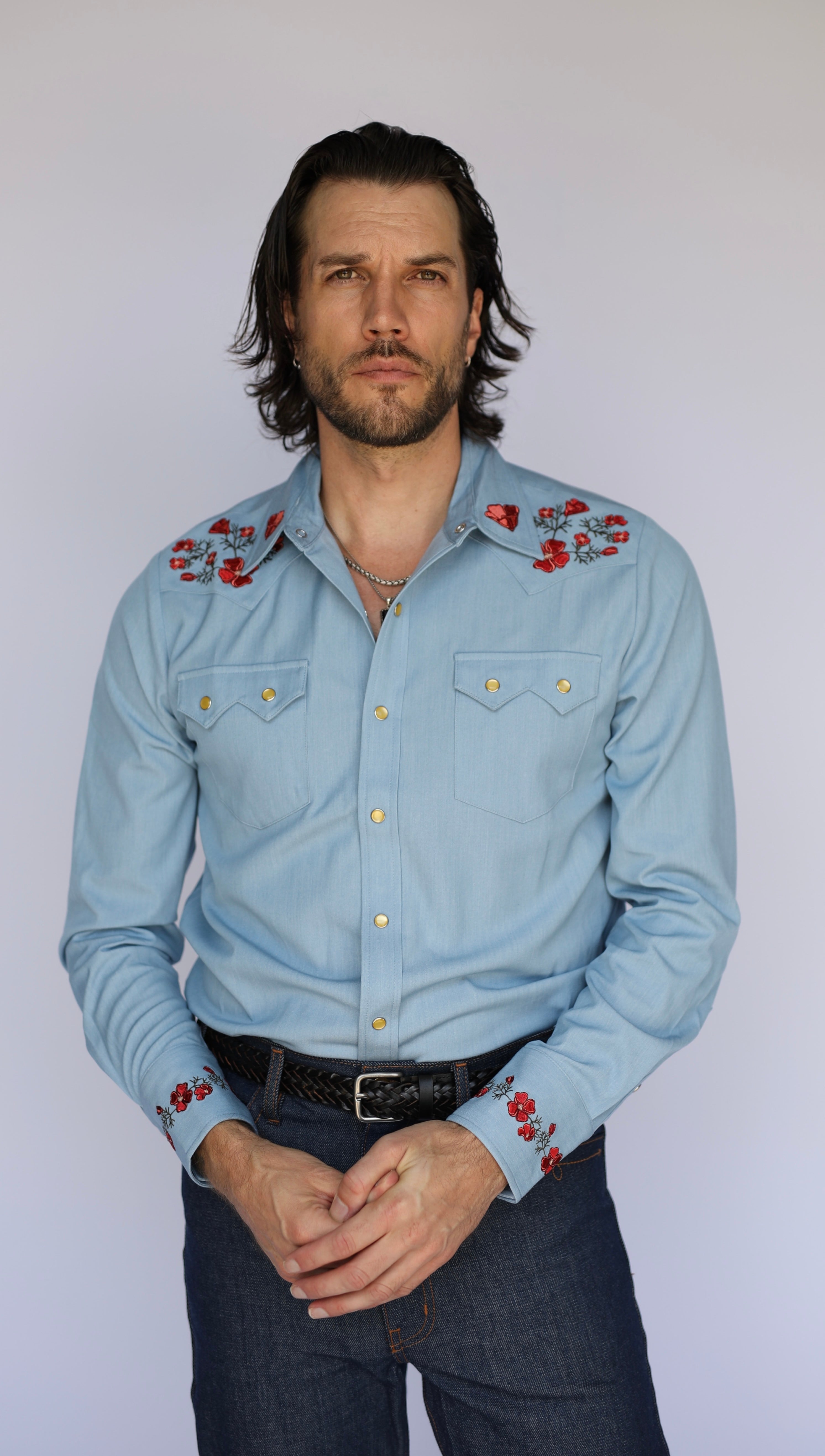 A man wearing a light blue western-style long-sleeve button-up shirt with red floral embroidery on the shoulders and cuffs, paired with dark jeans and a black belt, standing against a plain light gray background.