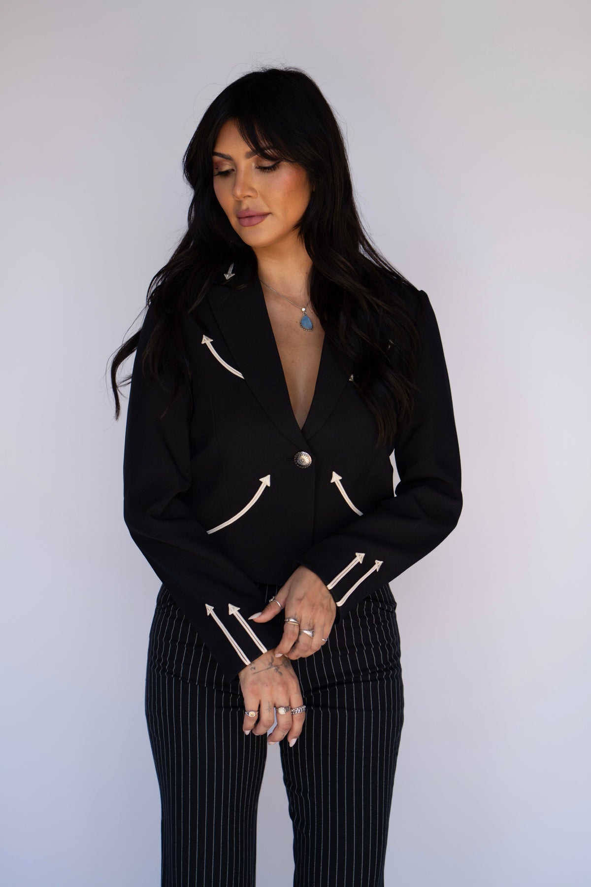 A woman modeling a black double-breasted blazer with white embroidered arrow details and matching black pinstriped trousers, standing against a plain light gray background.