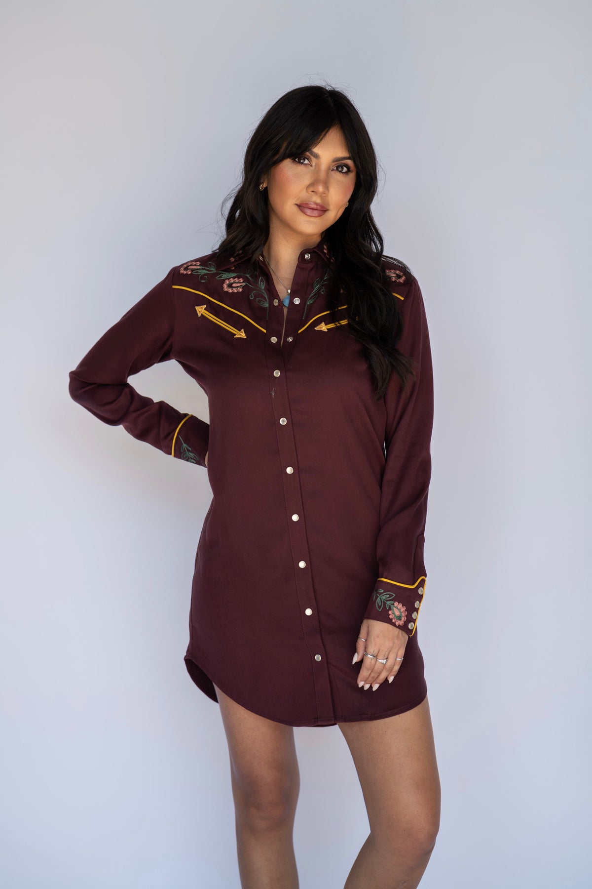 A woman modeling a long-sleeve western-style shirt dress in burgundy with embroidered details on the chest and cuffs, posed against a plain light gray background.