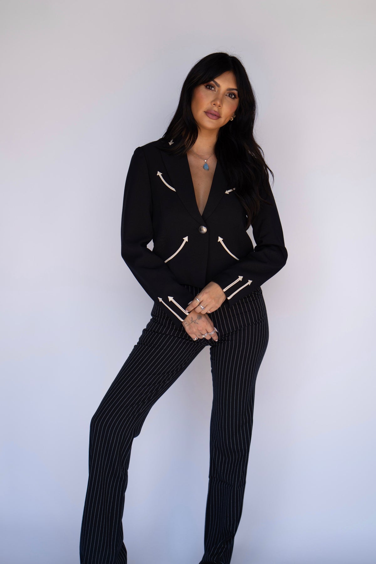 A woman modeling a black two-piece pantsuit with pinstripes and gold-tone hardware, standing against a plain light gray studio background.