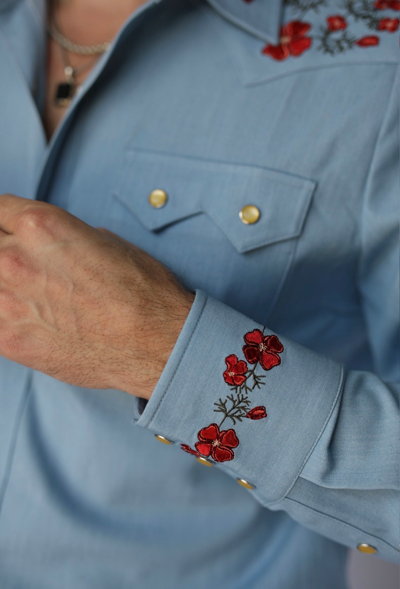 Close-up of a light blue western-style shirt worn by a model, featuring red floral embroidery on the chest and cuffs with gold snap buttons.