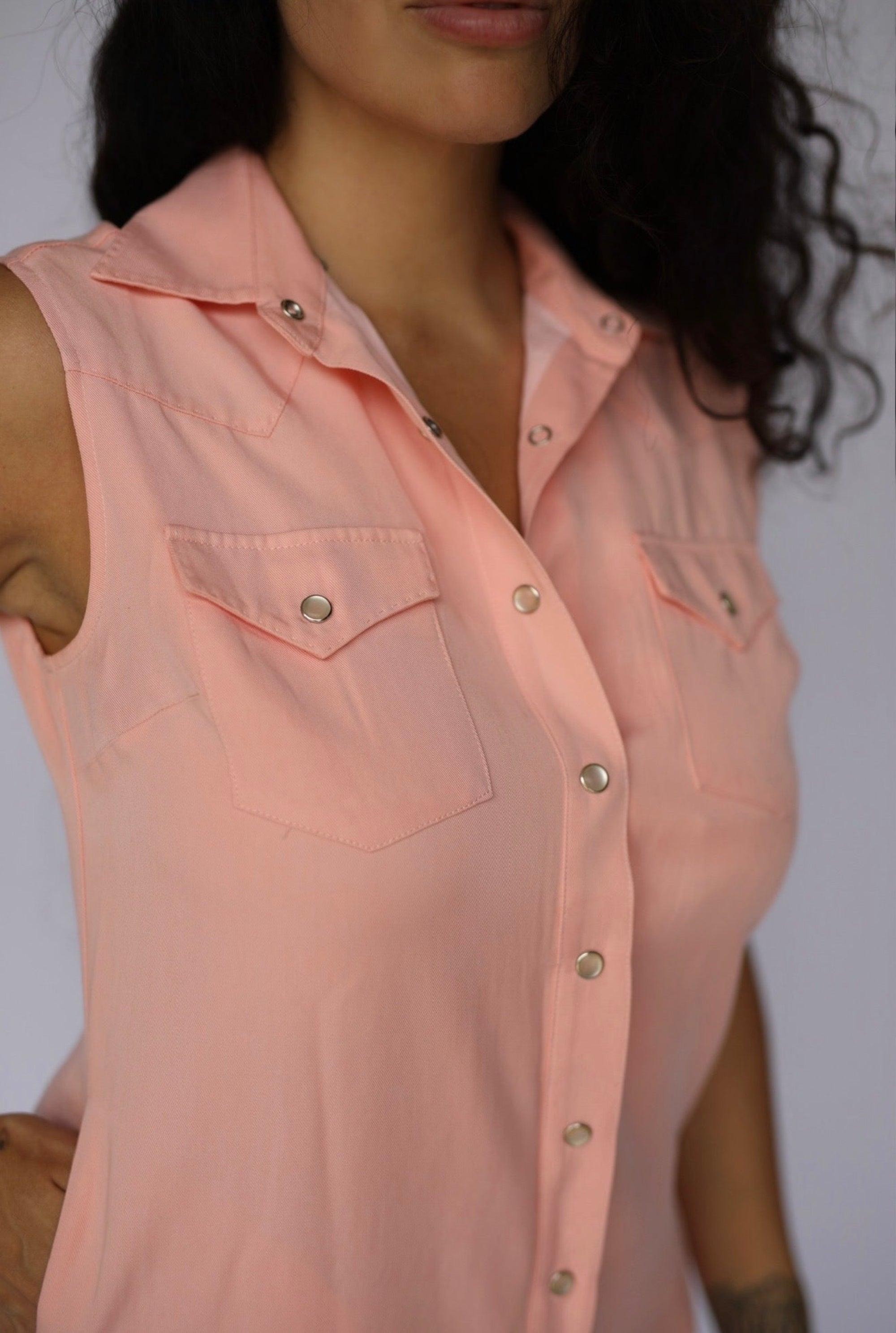 A close-up of a woman wearing a sleeveless, button-front collared top in a soft peach-pink color, with two chest pockets and silver-toned snap buttons.