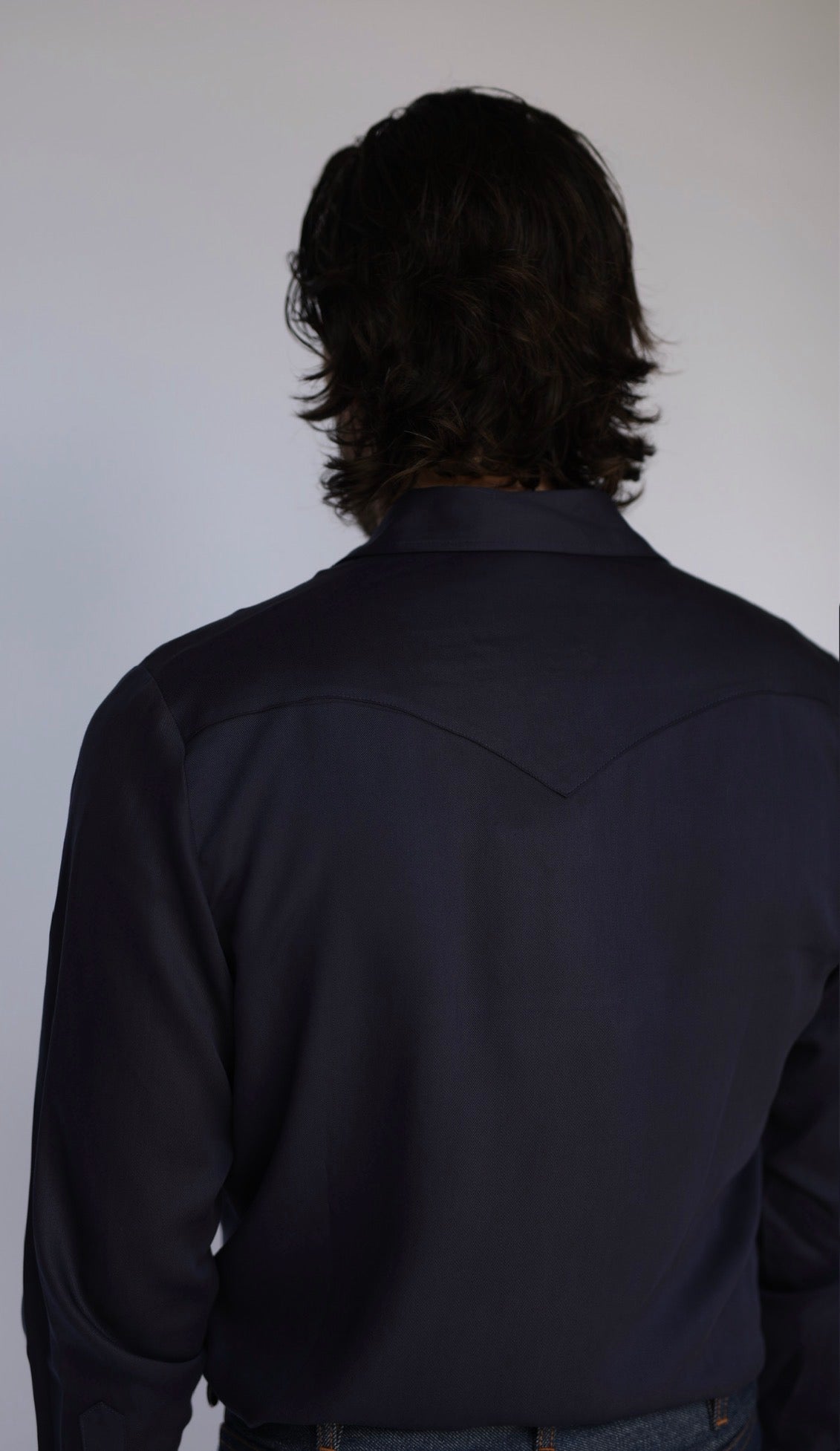Back view of a person with long dark hair wearing a dark navy long-sleeve western-style shirt against a plain light gray background.